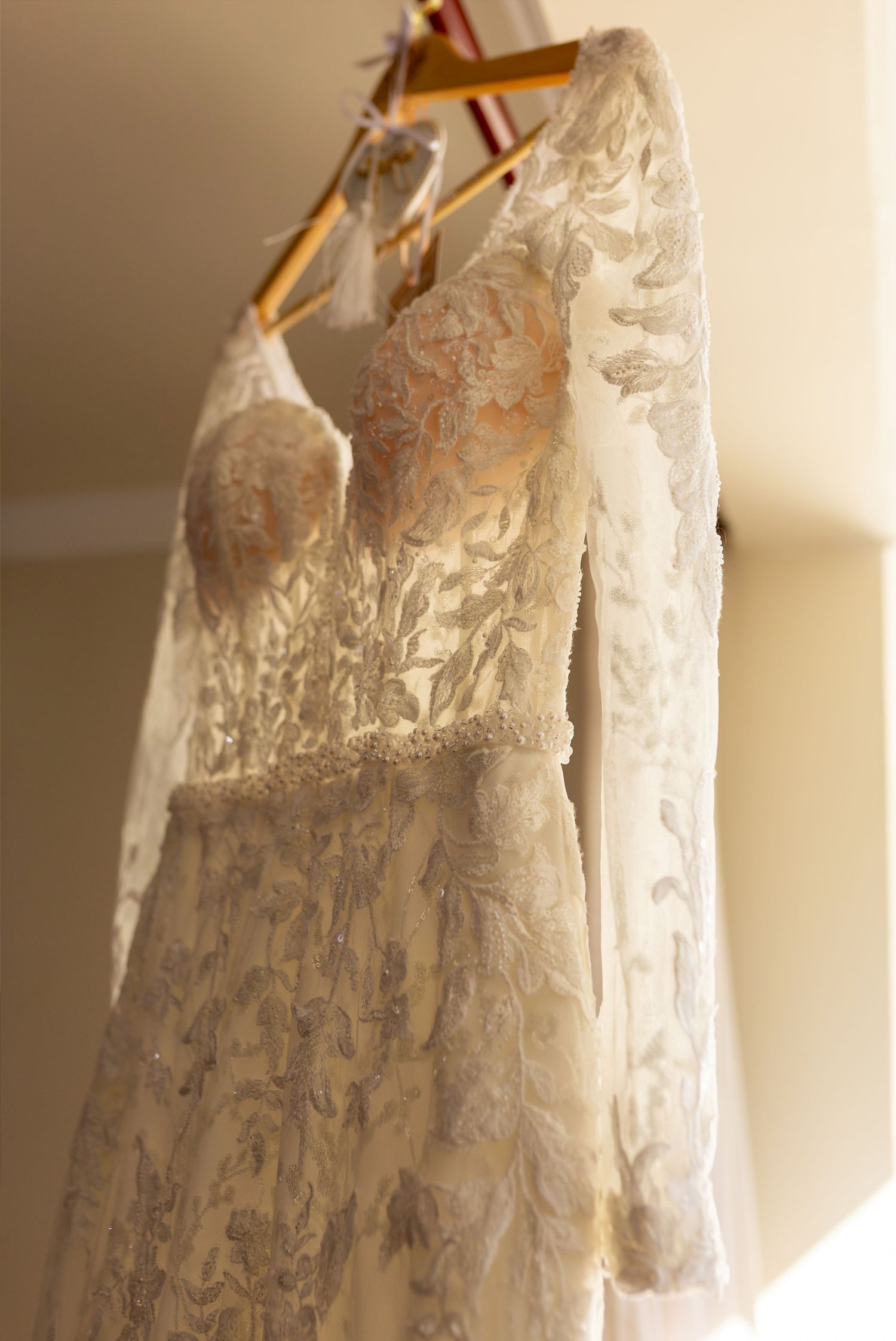 Elegant lace wedding dress with long sleeves hanging on a wooden hanger in soft warm light.