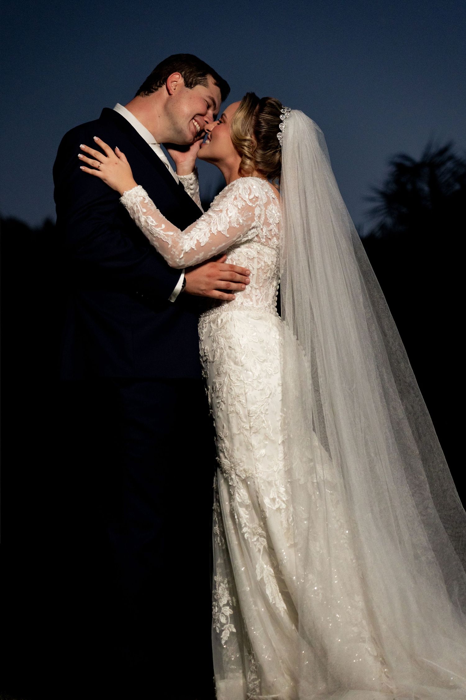 Bride and groom share a romantic kiss at dusk, bride in lace long-sleeve gown with veil, groom in navy suit.