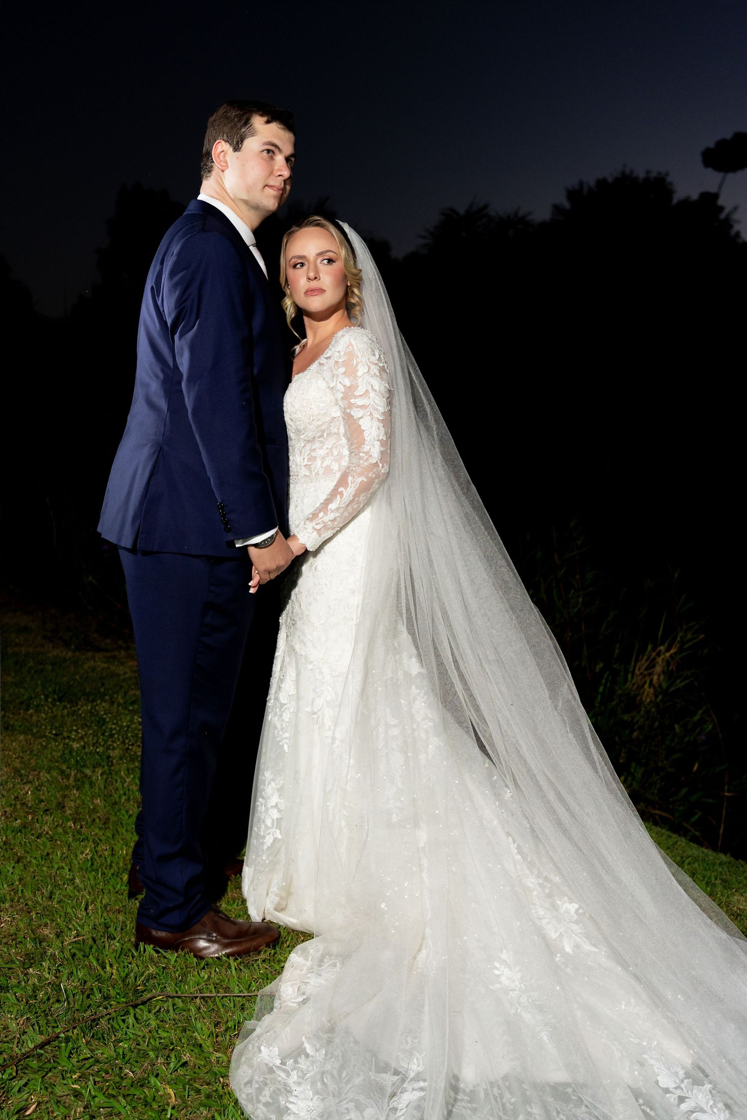Bride in lace gown with veil and groom in navy suit hold hands outdoors at night during wedding portrait.