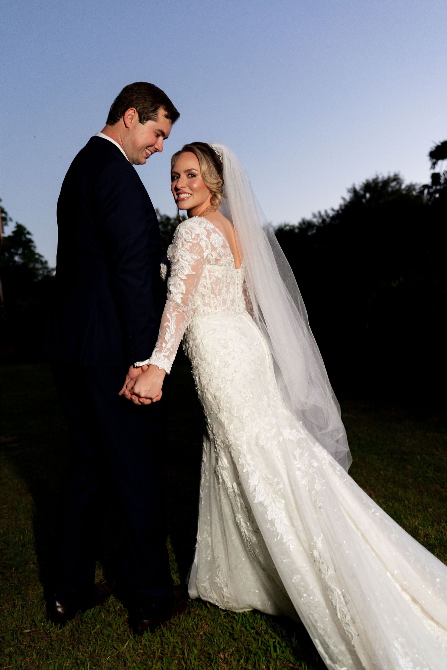 Bride in lace mermaid gown with veil holds hands with groom in navy tuxedo outdoors at dusk.