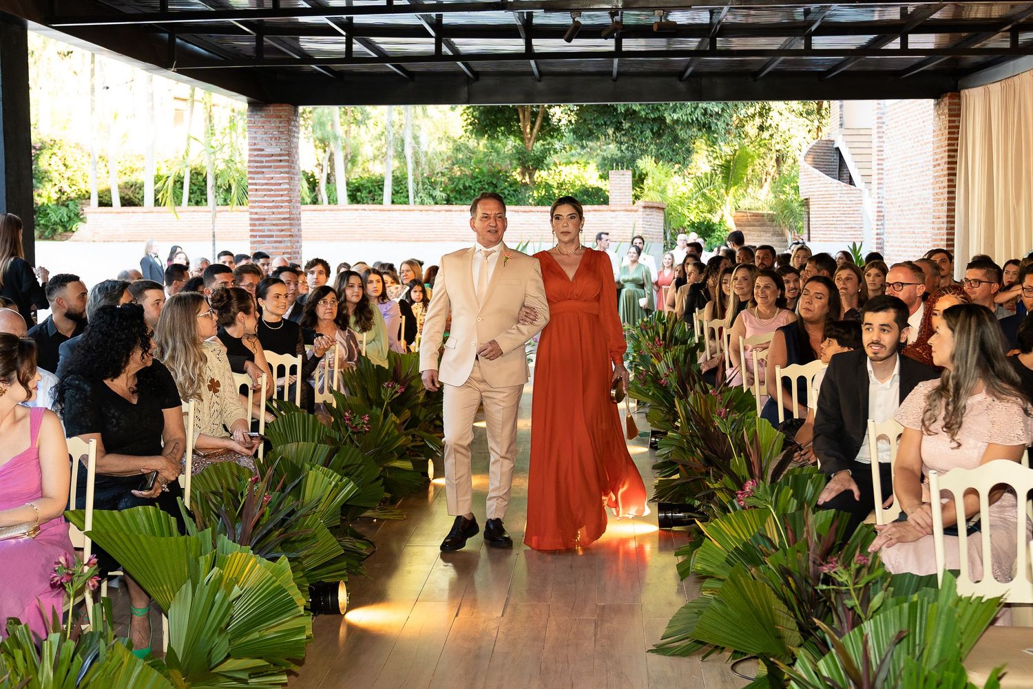 Couple walks down floral-lined wedding aisle outdoors, guests seated on both sides in elegant venue.