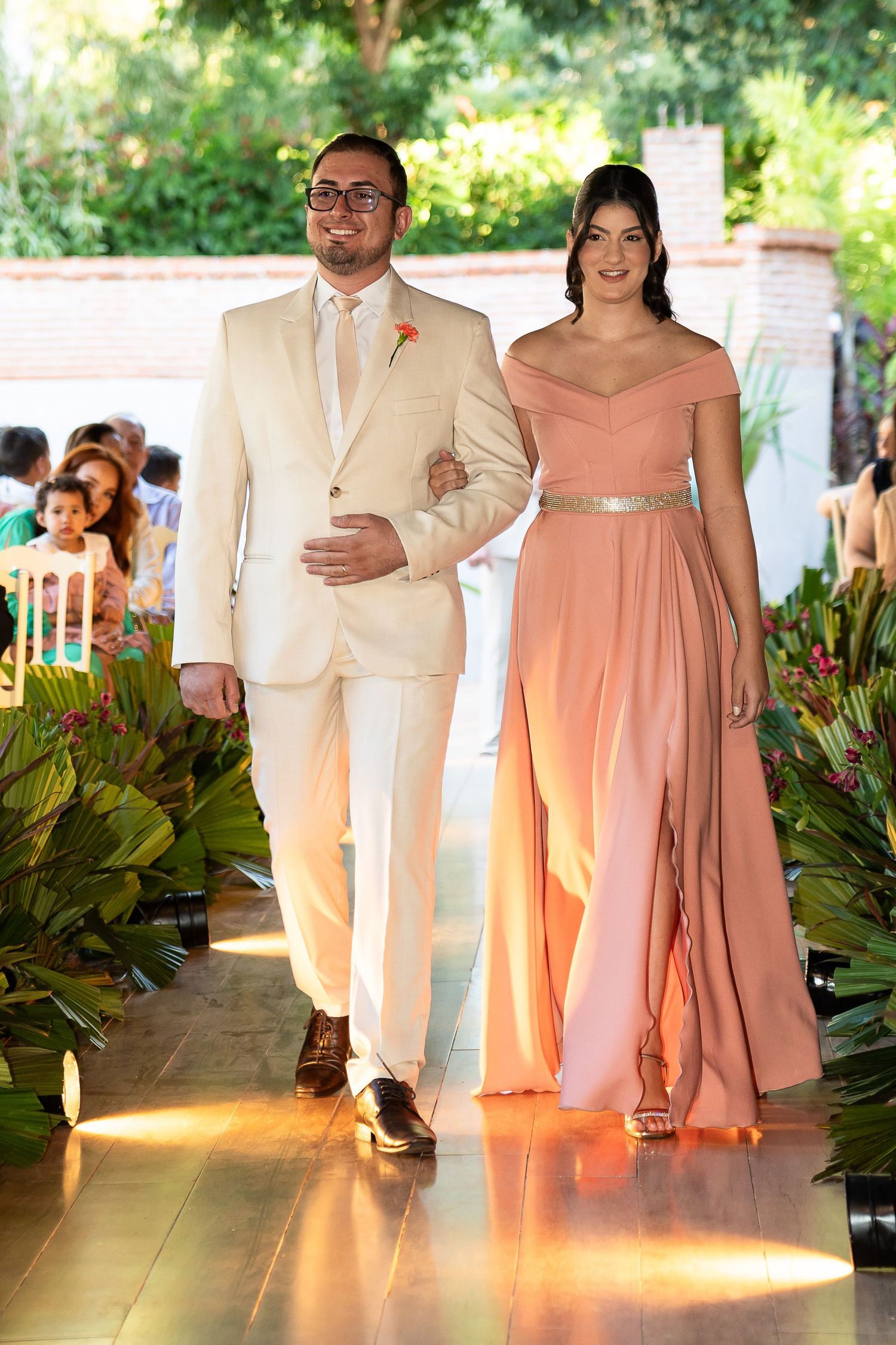 A man in a beige suit and woman in a peach gown walk down a floral wedding aisle together.