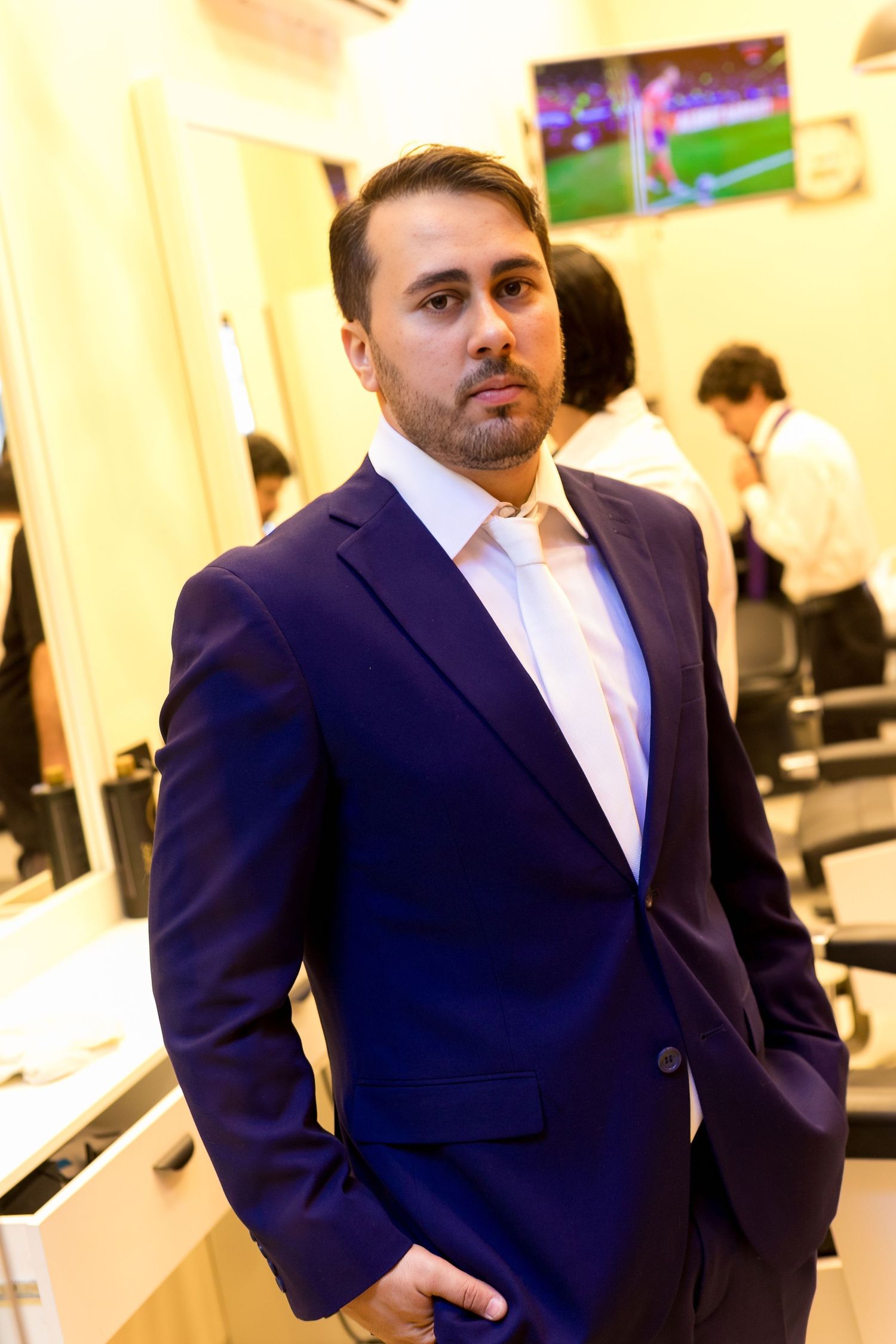 A well-dressed man in a navy blue suit poses confidently inside a modern barbershop with a TV screen in the background.