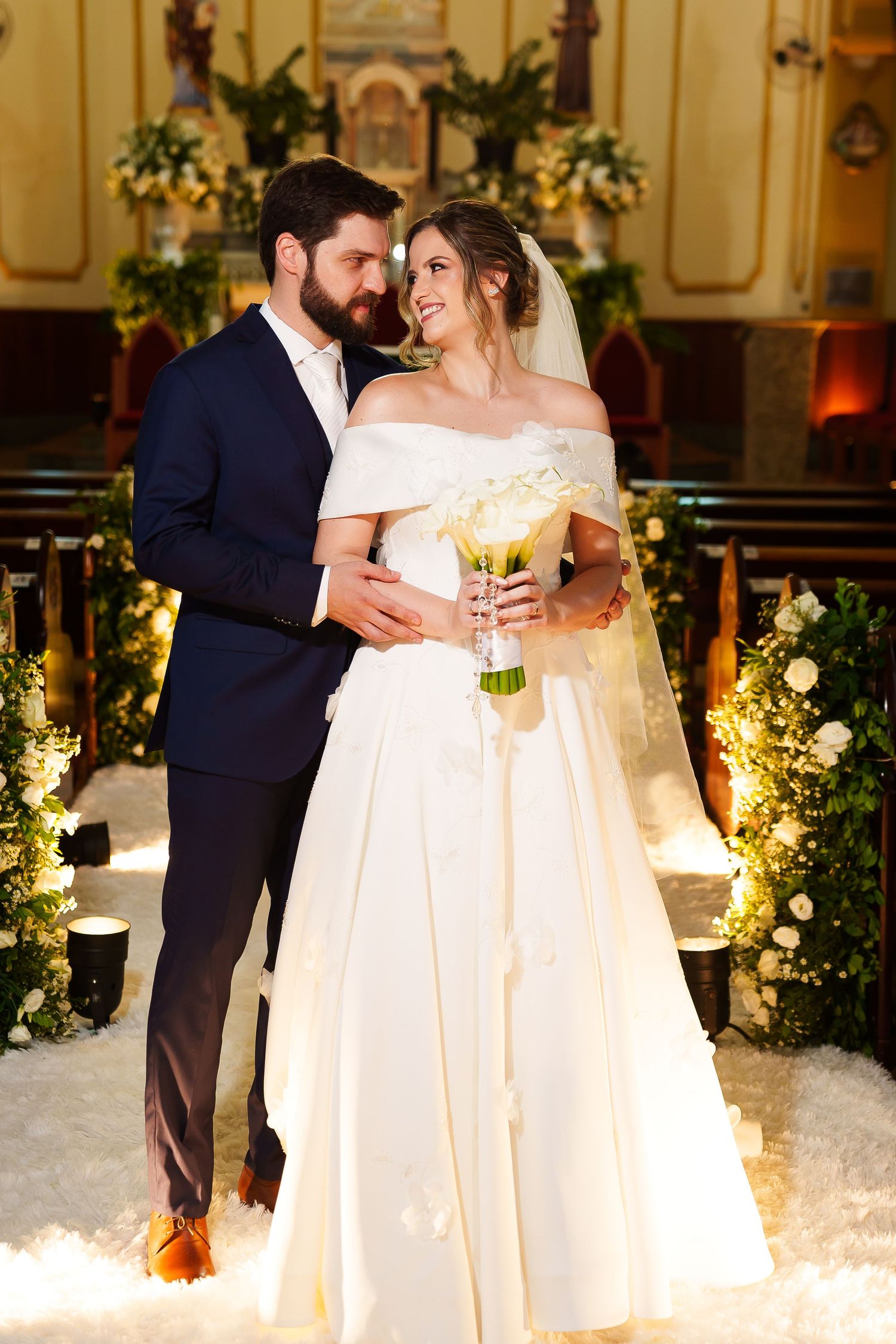 Bride and groom share a romantic moment at church altar, bride holding white bouquet in elegant off-shoulder gown.