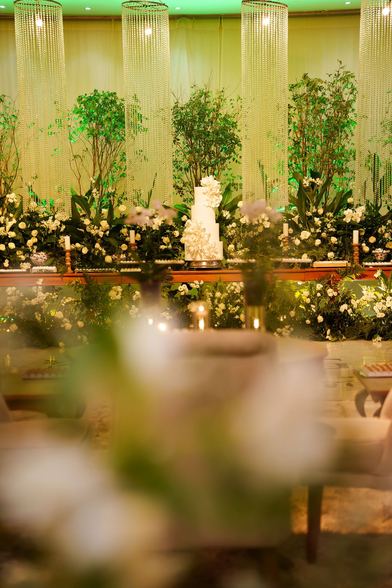 Elegant wedding reception with tiered white cake, lush greenery, white floral arrangements, and crystal chandeliers in a softly lit venue.