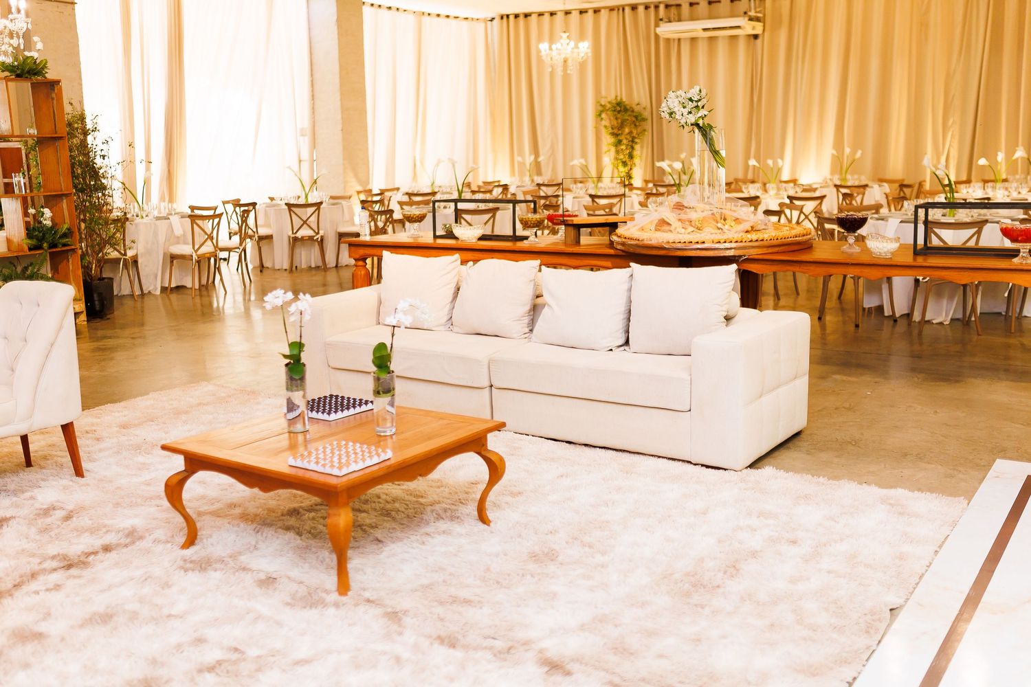 Elegant wedding reception venue with white sofas, wooden coffee table, and decorated dining tables under warm lighting.