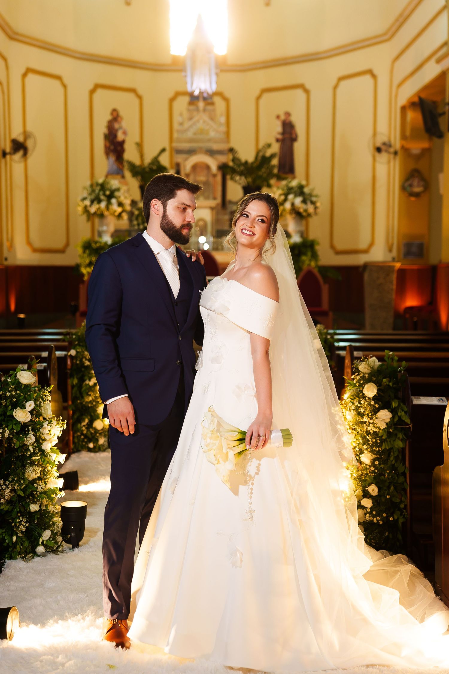 Bride in white off-shoulder gown and groom in navy suit pose together inside a flower-adorned church.