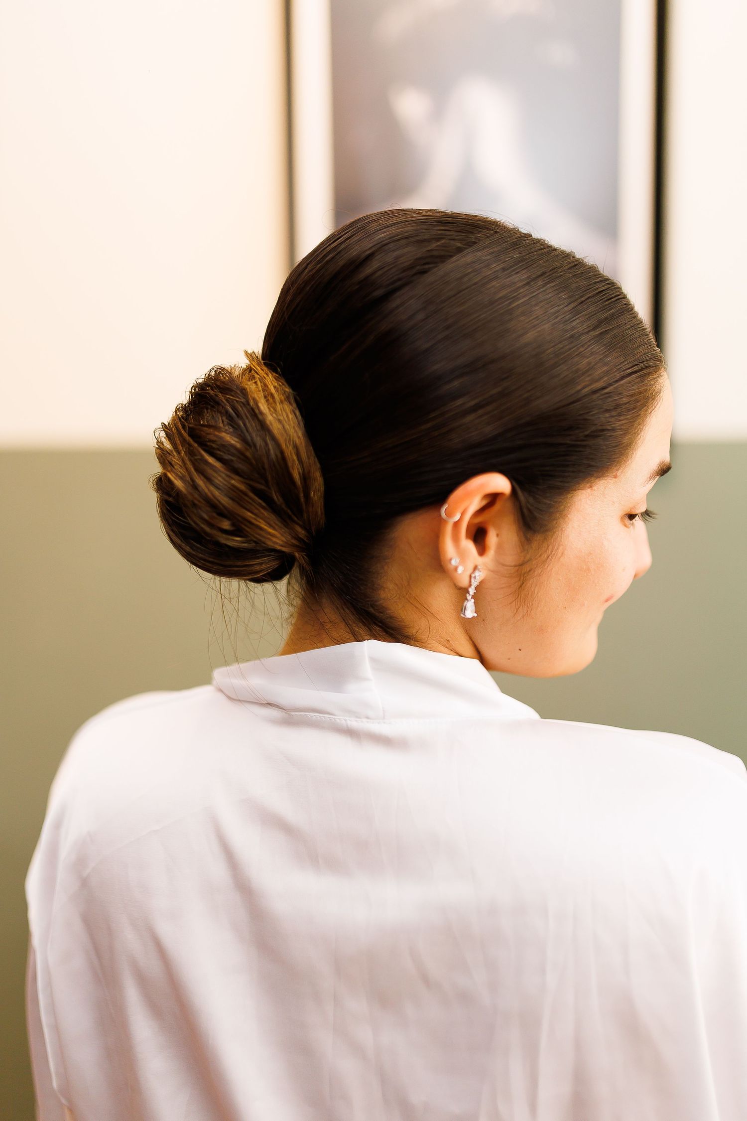 Woman with sleek dark hair in a low bun, wearing a white blouse and elegant drop earring, viewed from behind.
