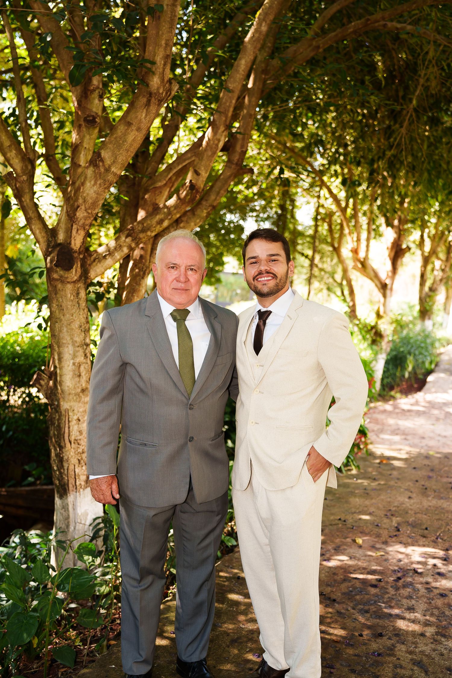 Two men in suits pose outdoors at a wedding, surrounded by lush green trees on a sunny day.