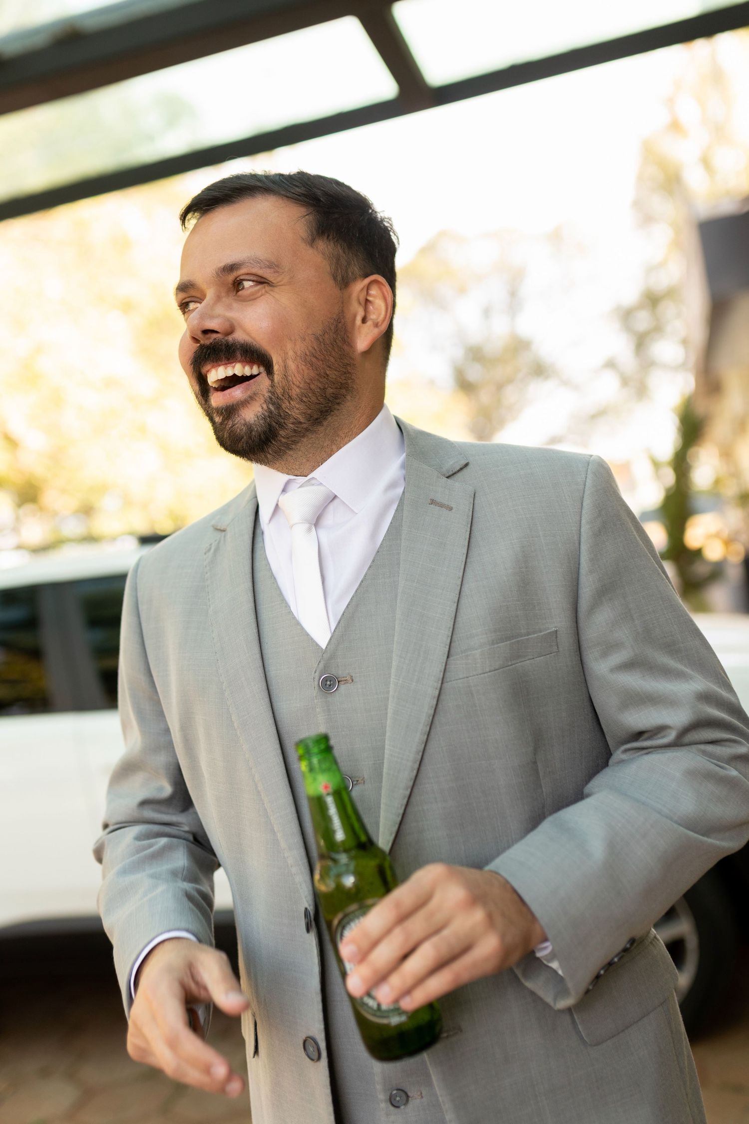 A laughing bearded man in a gray suit holds a green beer bottle outdoors near a white vehicle.