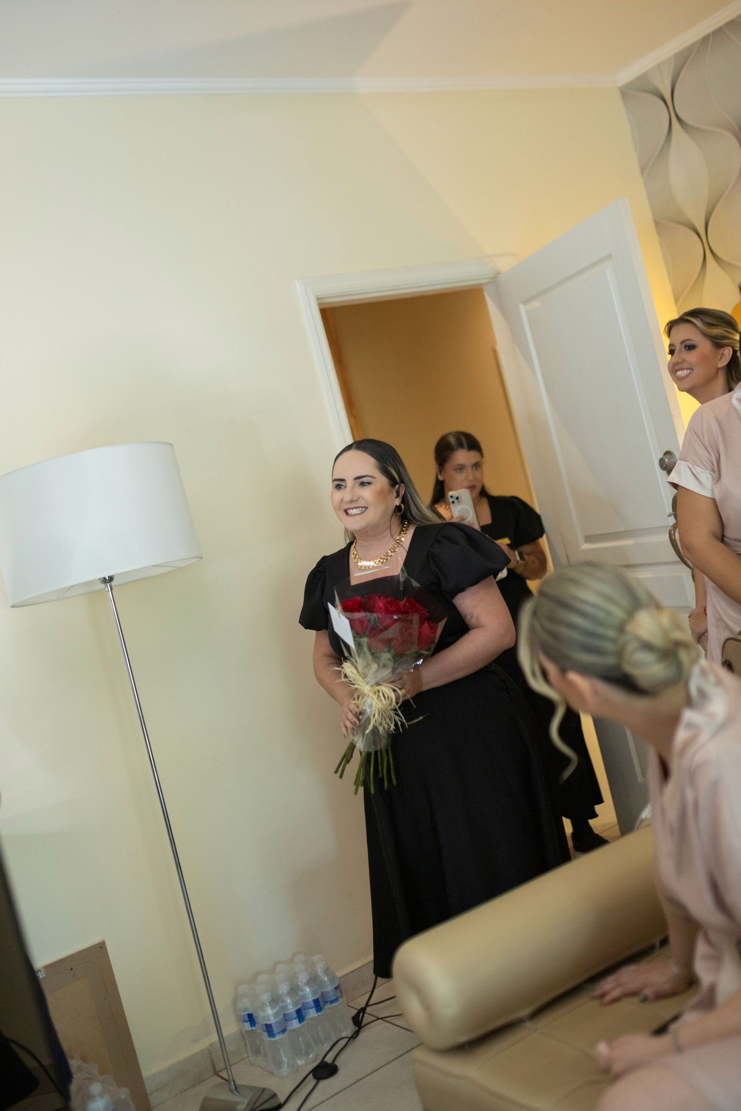Woman in black dress holding bouquet smiles in decorated room with bridesmaids during wedding preparations.