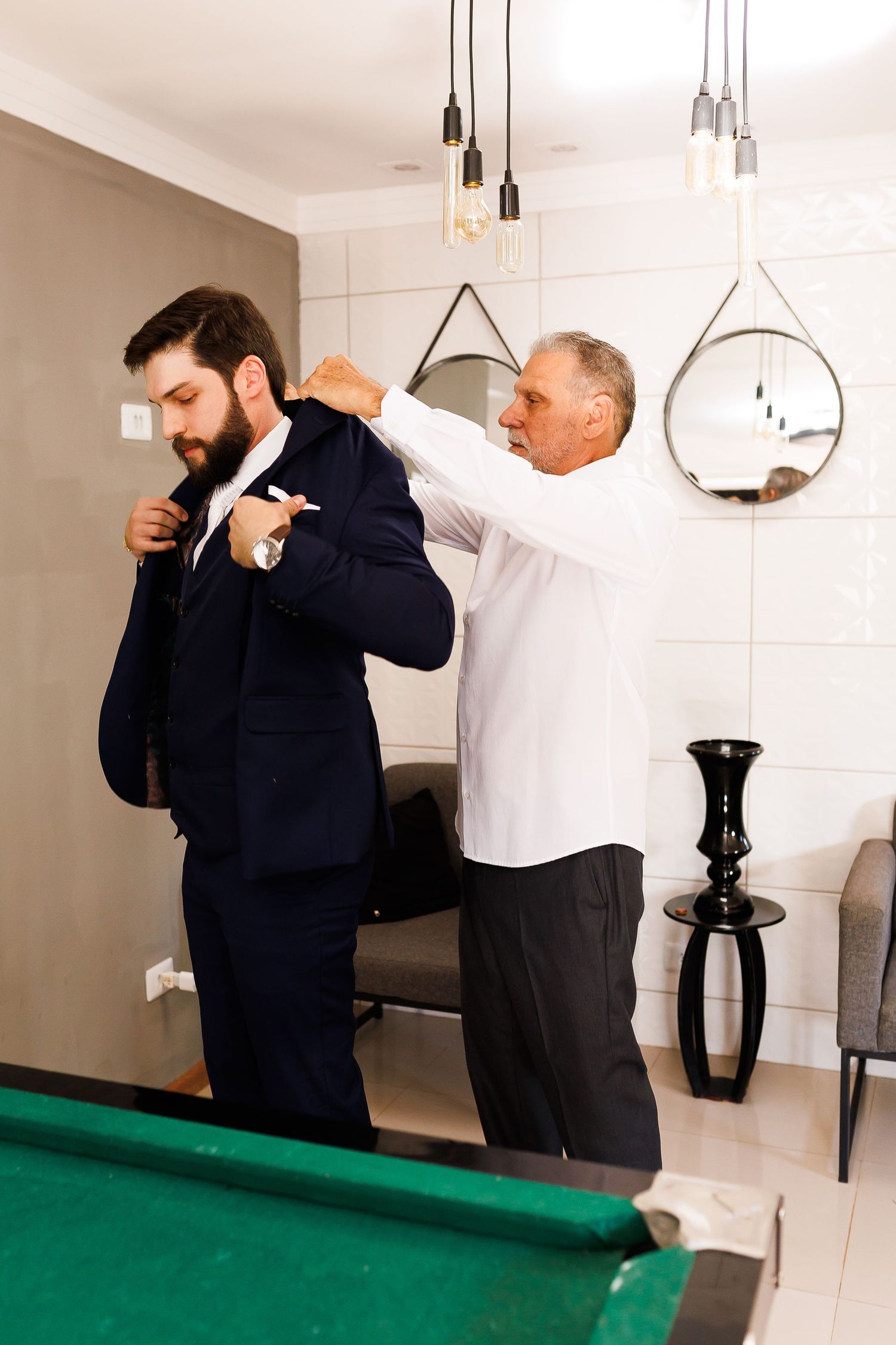 Older man helping a young bearded groom put on a navy suit jacket in a modern room with a pool table.