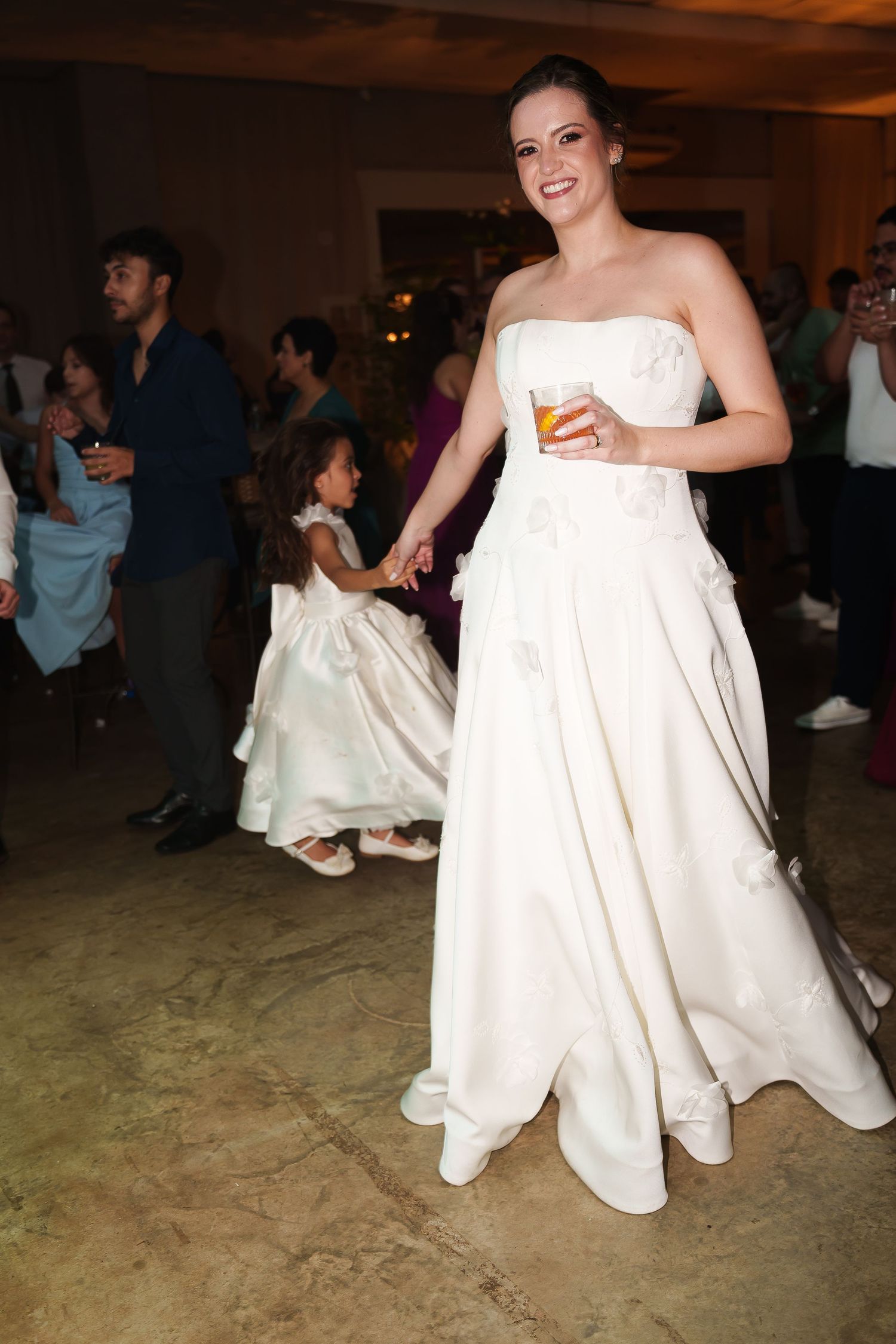 Bride in strapless white gown holds a drink while dancing with a flower girl at a wedding reception.