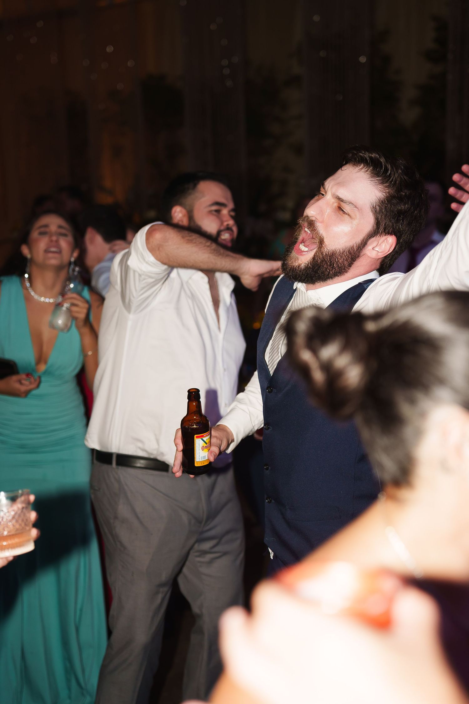 Two men dancing energetically at a wedding reception, one holding a beer bottle, surrounded by guests in formal attire.