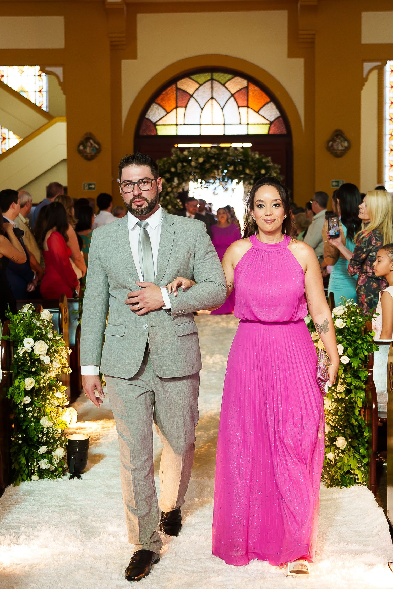 Couple walking down church aisle, man in gray suit, woman in pink gown, floral decor and guests in background.