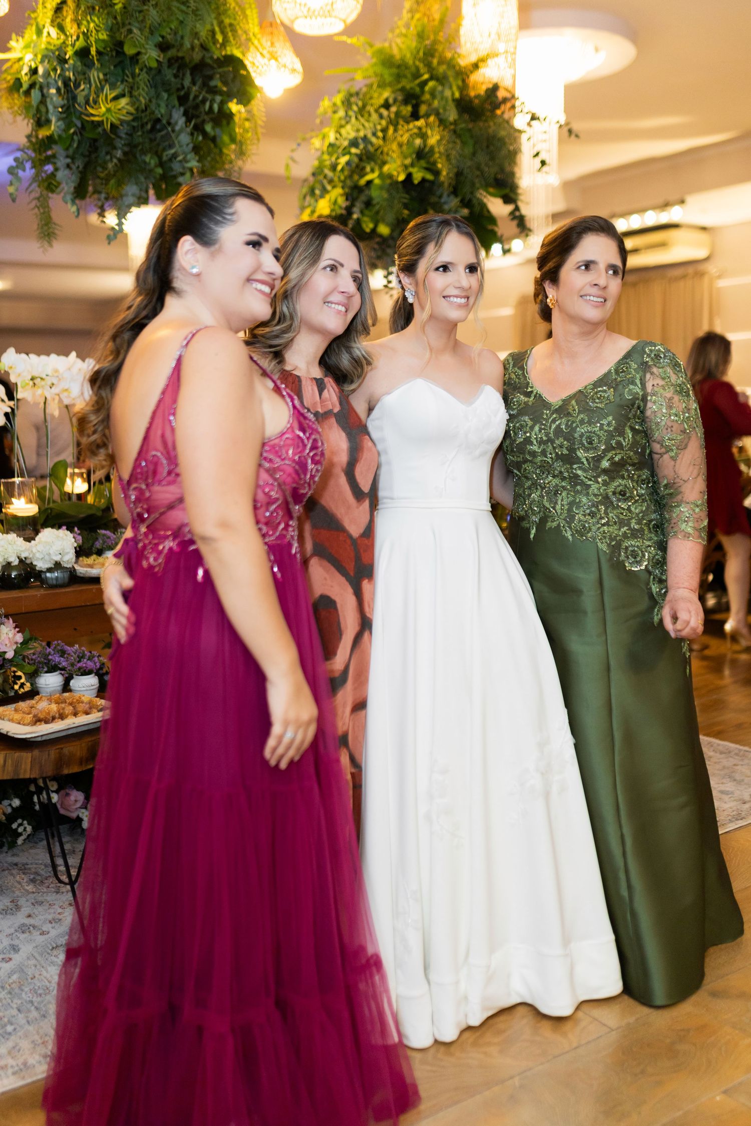 Bride in white gown poses with three women in elegant burgundy and green formal dresses at a wedding reception.
