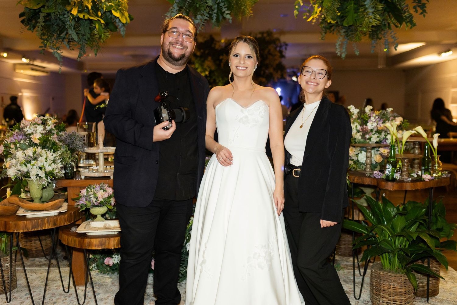 Bride in white gown poses with two guests in black attire at an elegantly decorated wedding reception venue.