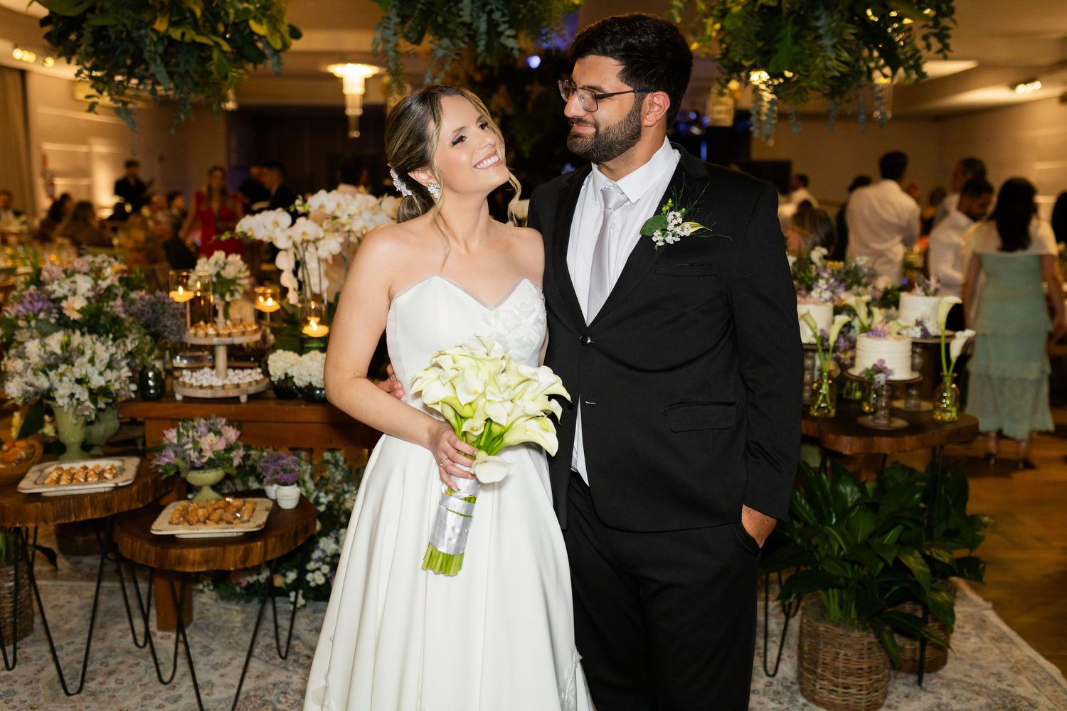 Bride and groom pose together at elegant wedding reception, bride holding white bouquet in bridal gown, groom in black suit.