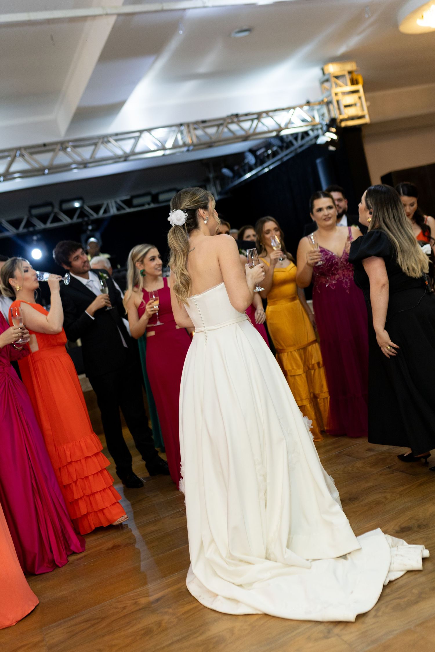 Bride in white gown stands on dance floor surrounded by guests in colorful formal attire at wedding reception.