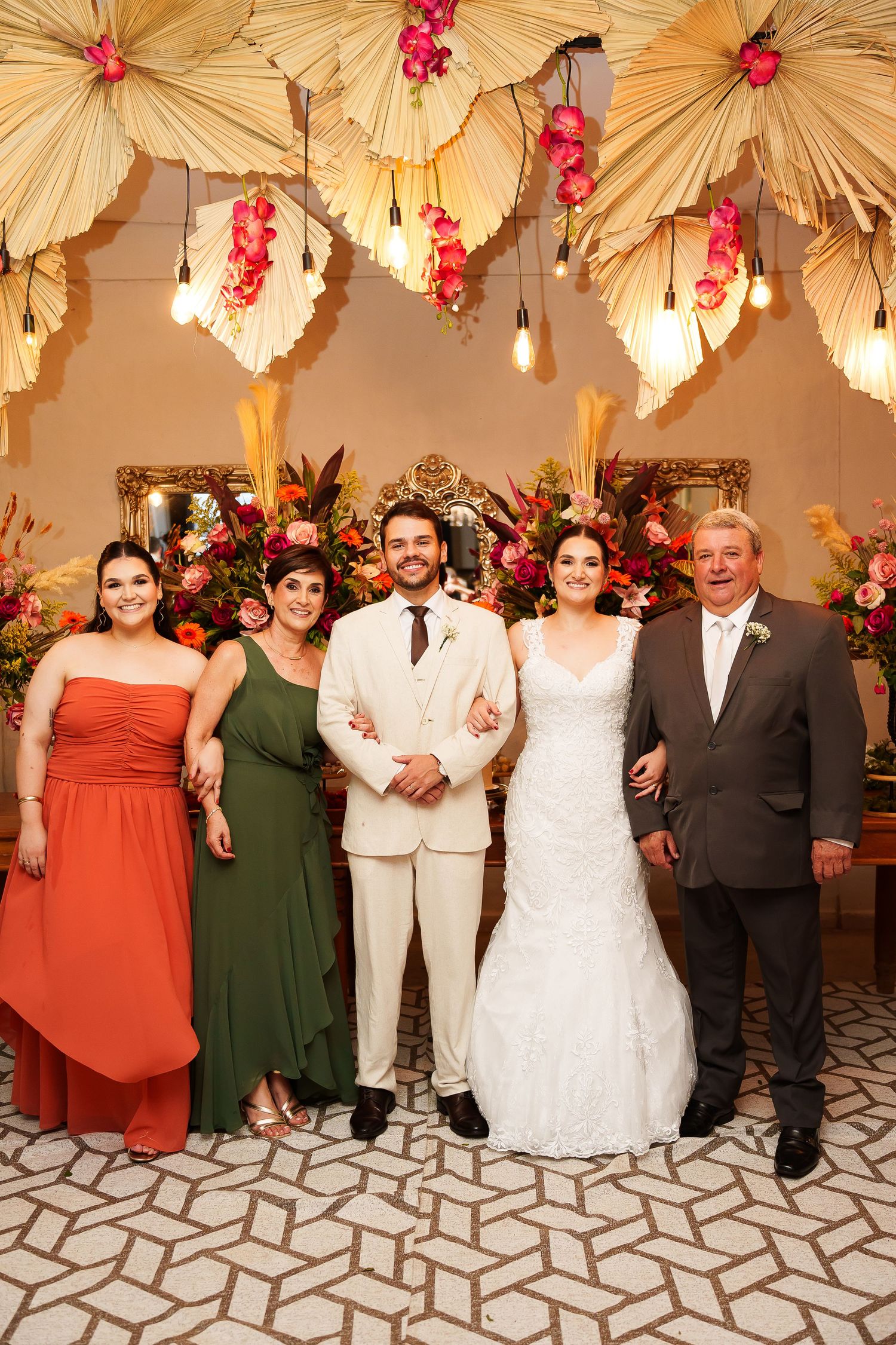 Wedding party poses before floral backdrop, bride in white gown, groom in cream suit, guests in orange and green dresses.