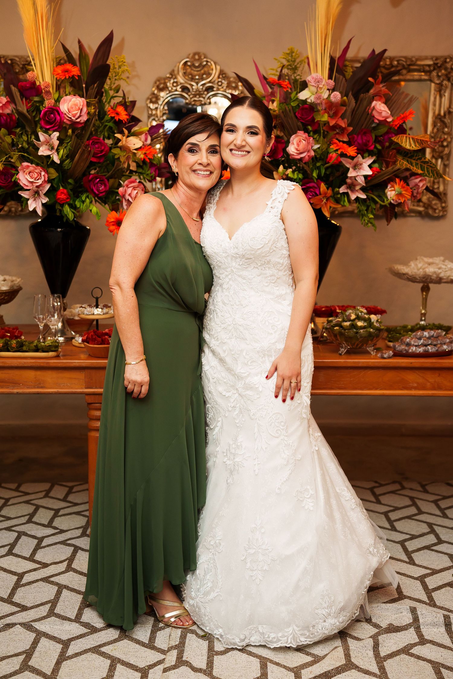 Bride in white lace gown poses with woman in green dress before floral backdrop at elegant wedding reception.