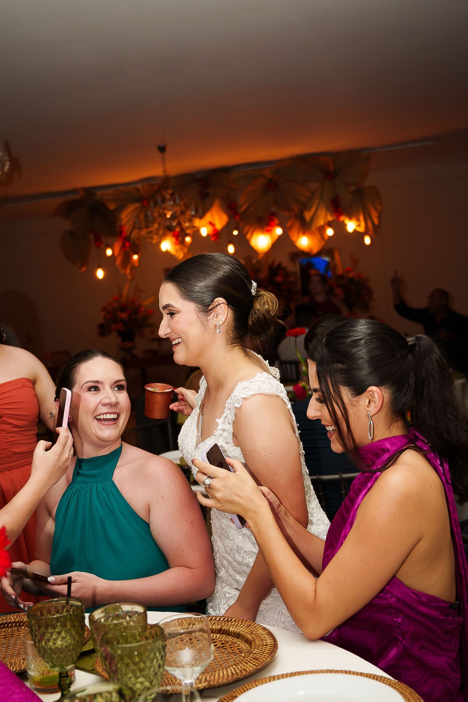 Bride in white lace gown laughing with bridesmaids in teal and purple at a warmly lit wedding reception.