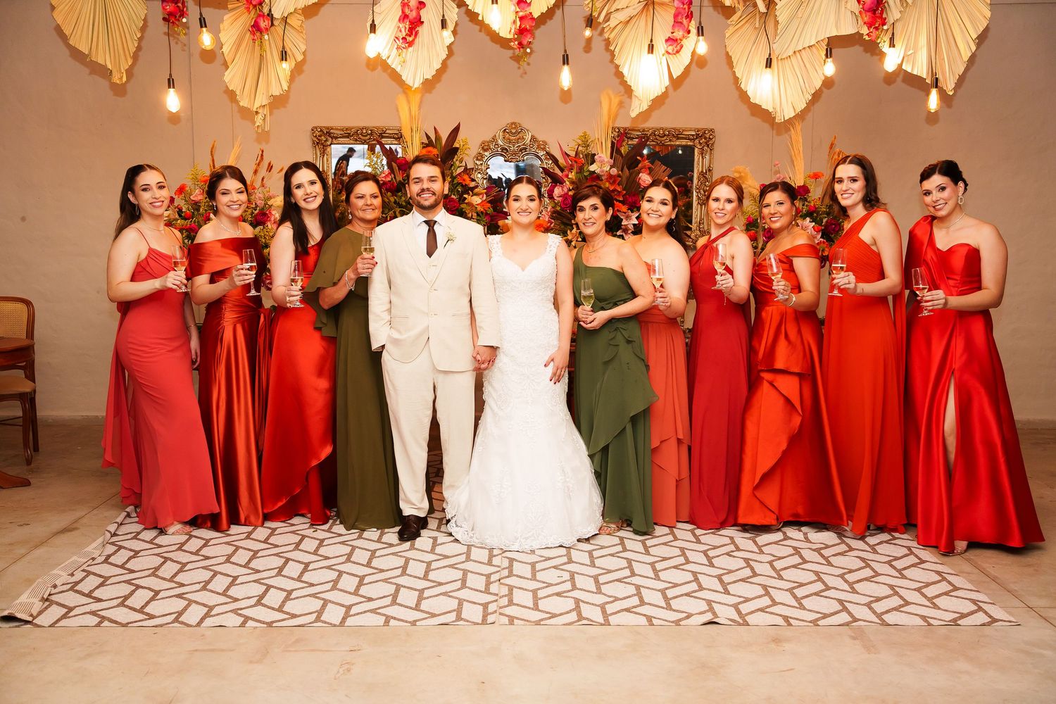 Wedding party poses together, bride in white gown with groom in beige suit, surrounded by bridesmaids in red and olive dresses.