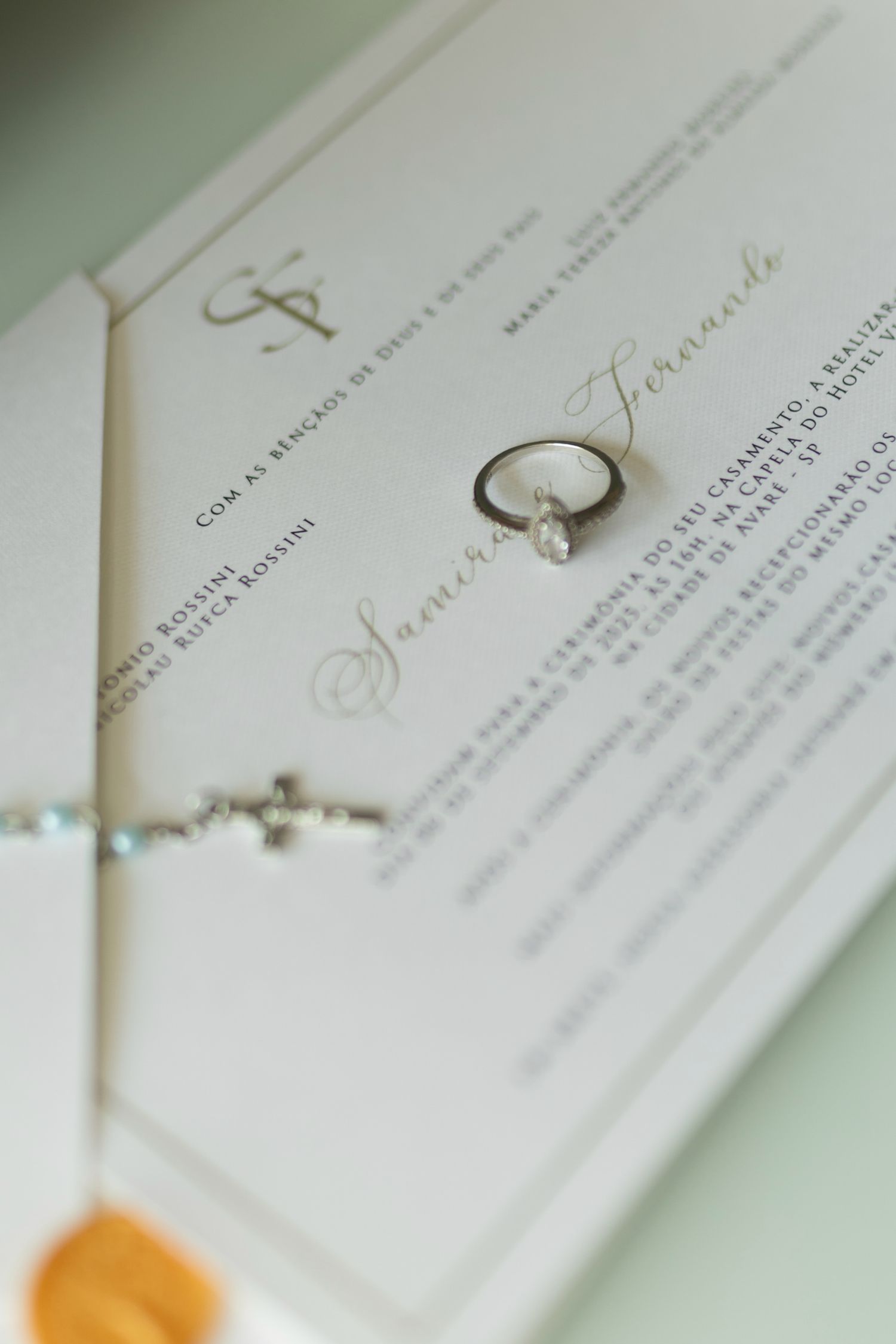 Wedding invitation with diamond engagement ring and cross bracelet on white paper with gold script text.