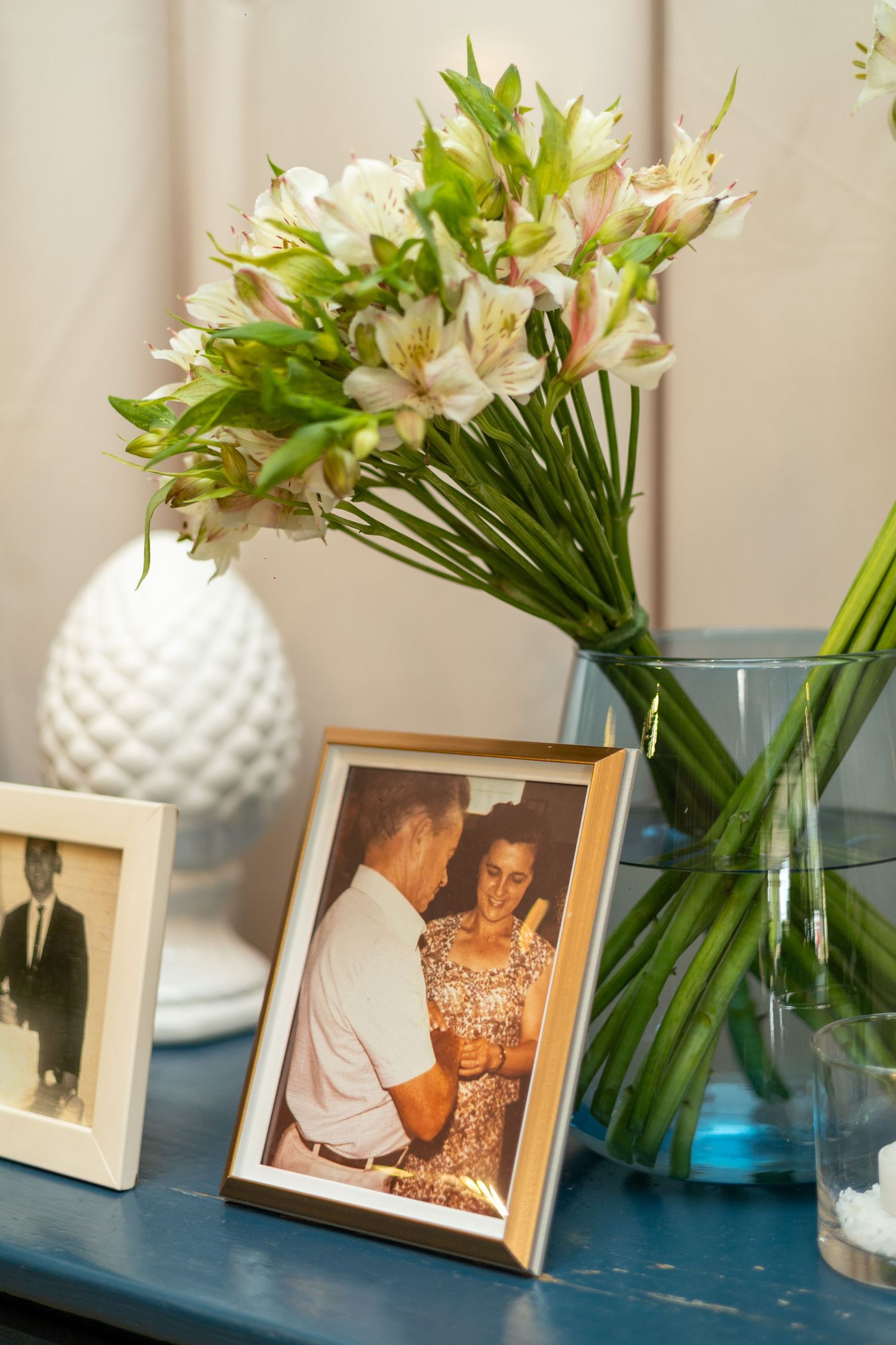 Framed family photos on a blue shelf with white alstroemeria flowers in a glass vase and decorative white vase.
