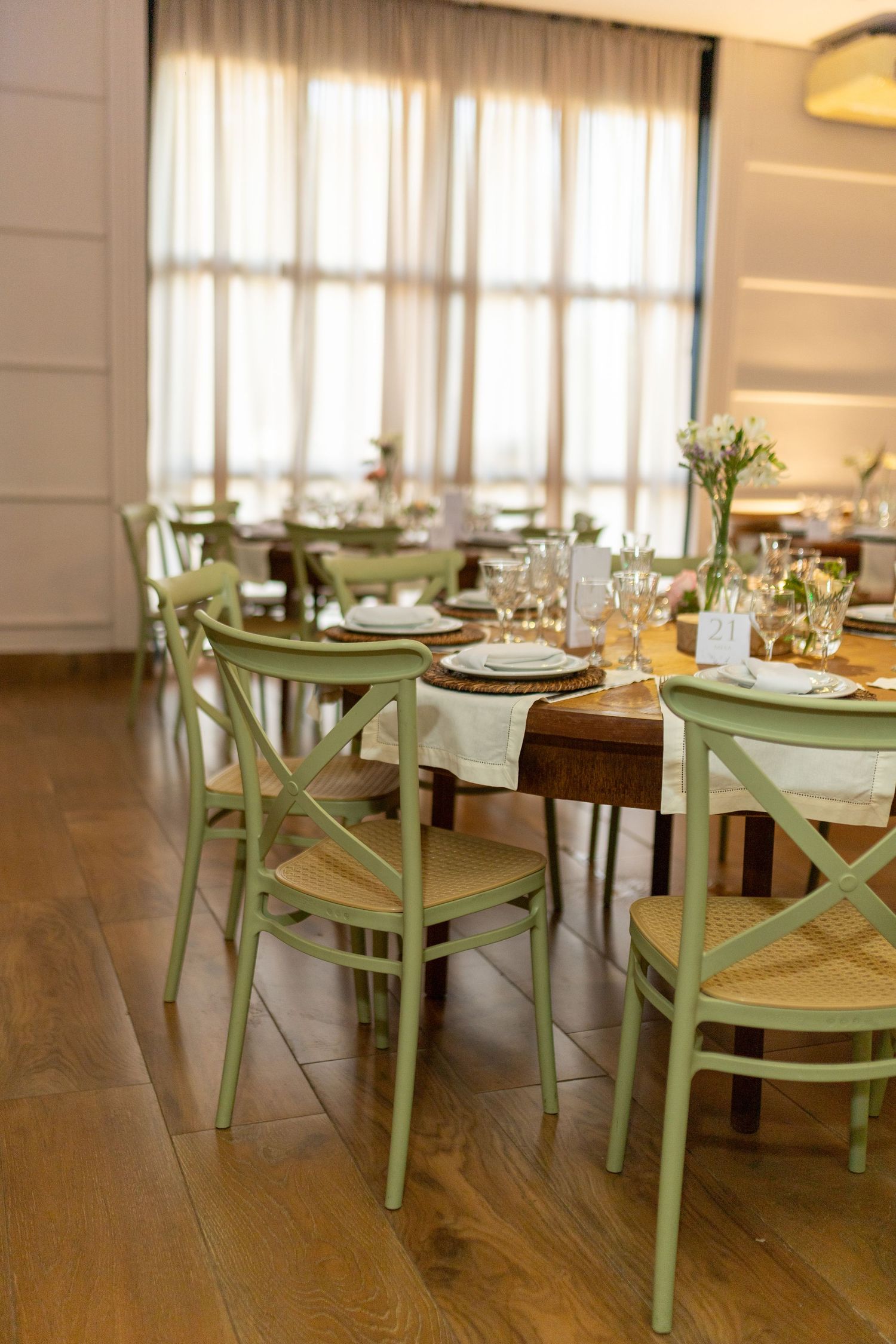 Elegant restaurant dining room with sage green cross-back chairs and wooden tables set with glassware and white plates.