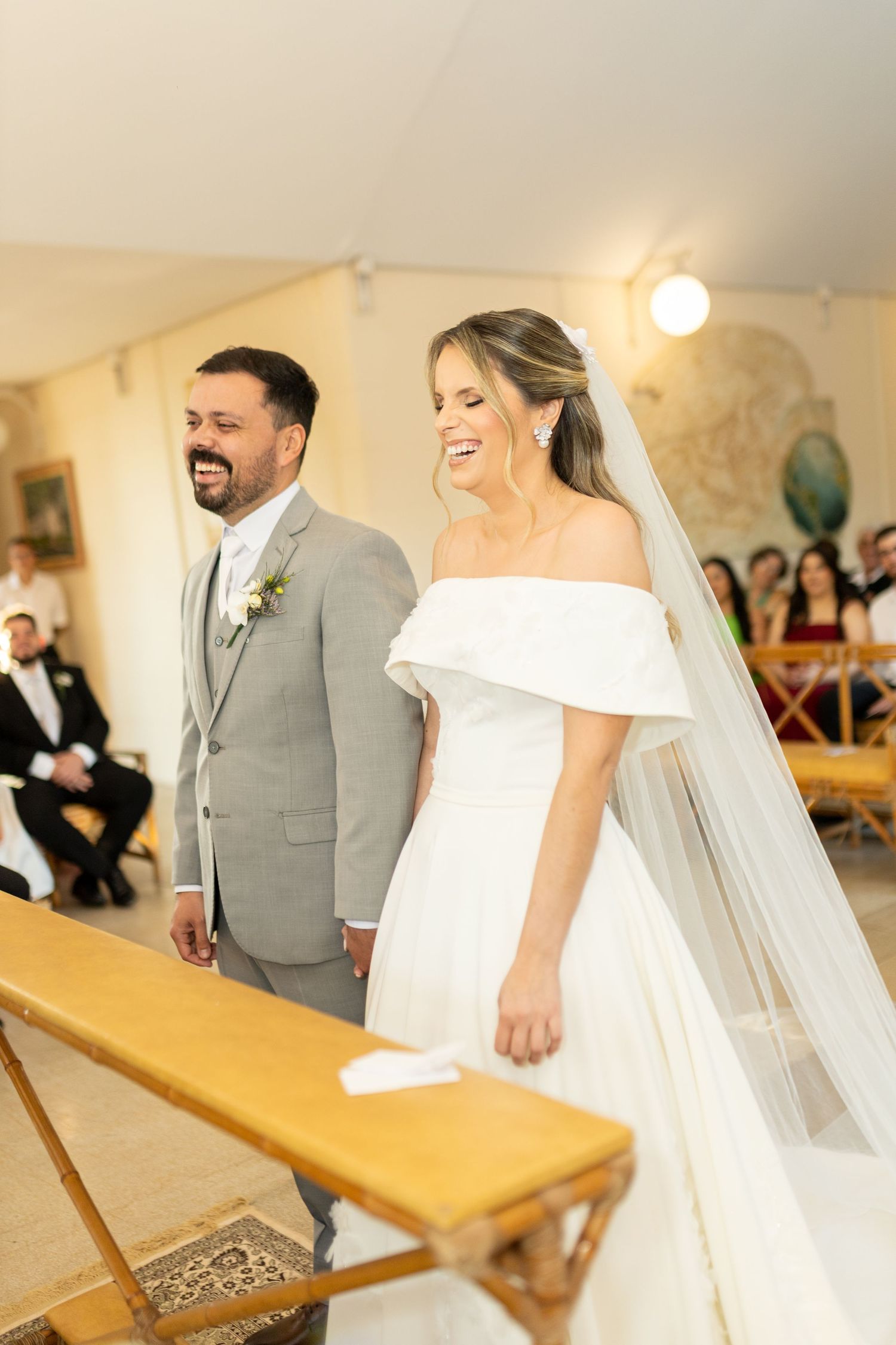 Bride in off-shoulder gown with veil and groom in gray suit smiling joyfully at wedding ceremony altar.