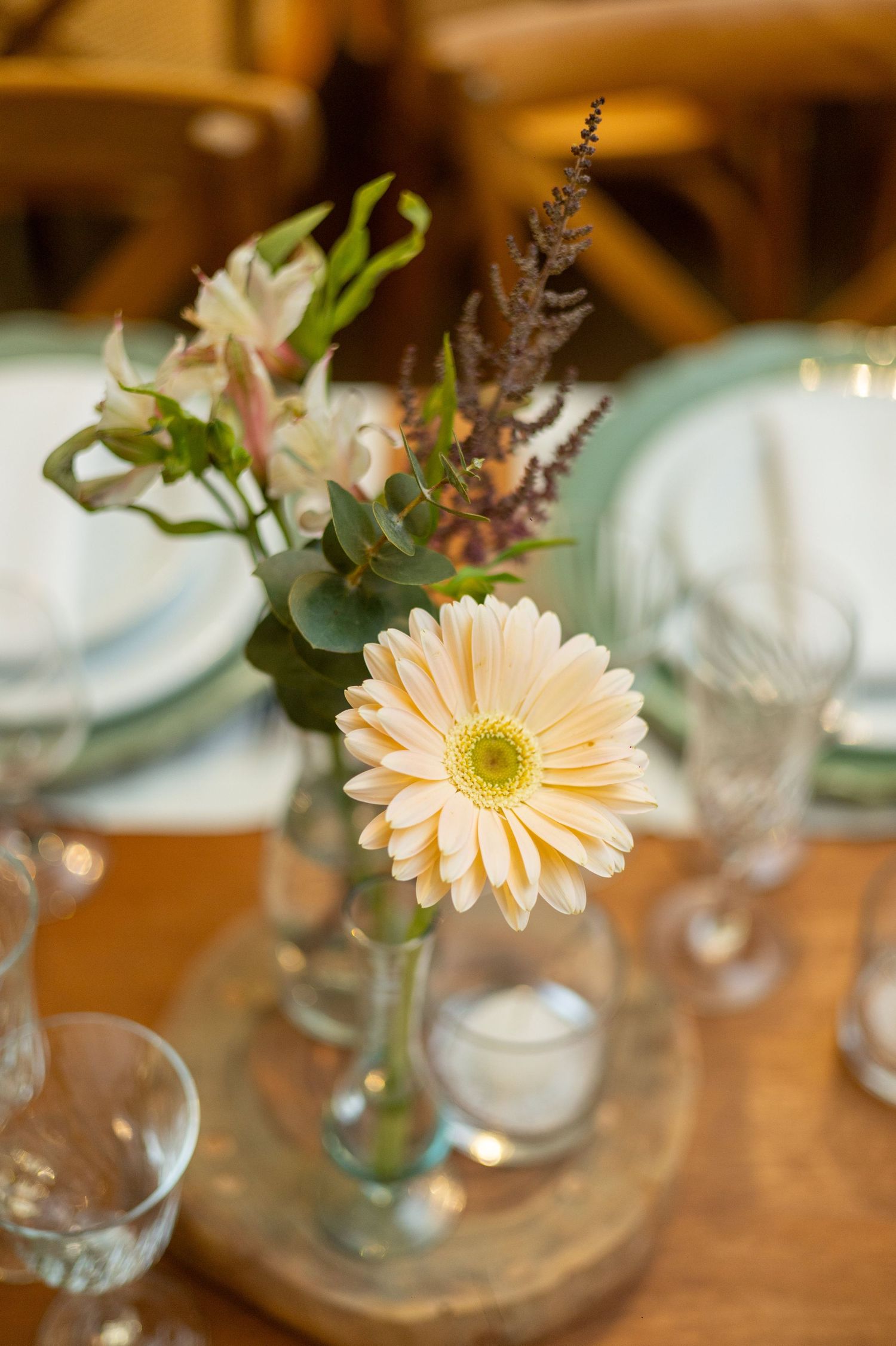 Elegant table centerpiece with a peach gerbera daisy in a glass vase, surrounded by mixed flowers and candles on wood.
