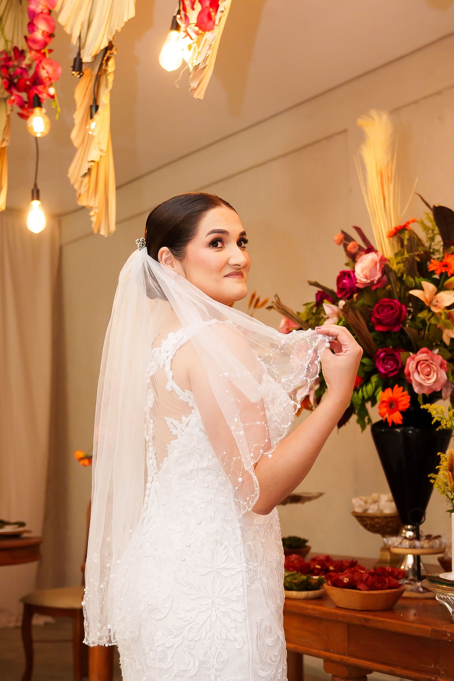 Elegant bride in white lace wedding gown and veil posing at reception table with colorful floral arrangements.