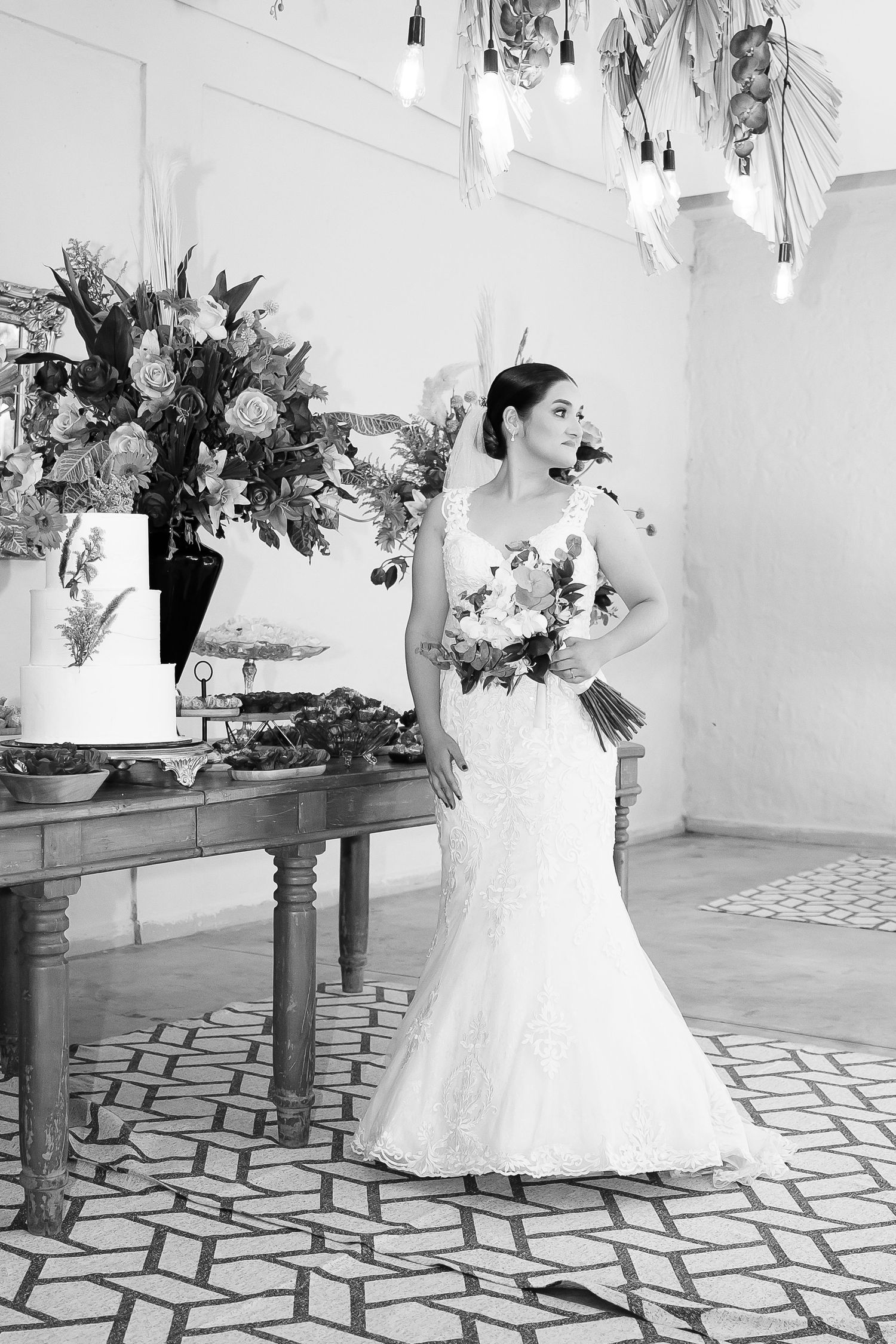 Bride in lace mermaid wedding gown holding bouquet stands beside floral arrangements and tiered cake in elegant venue.