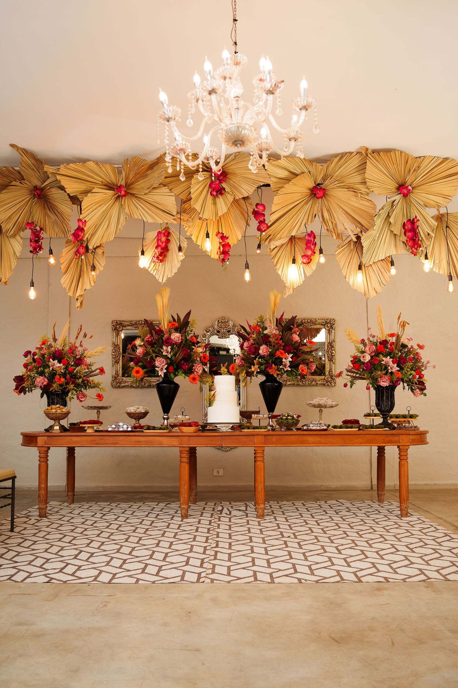 Elegant event table with floral arrangements, white cake, golden palm leaf decor, and crystal chandelier overhead.