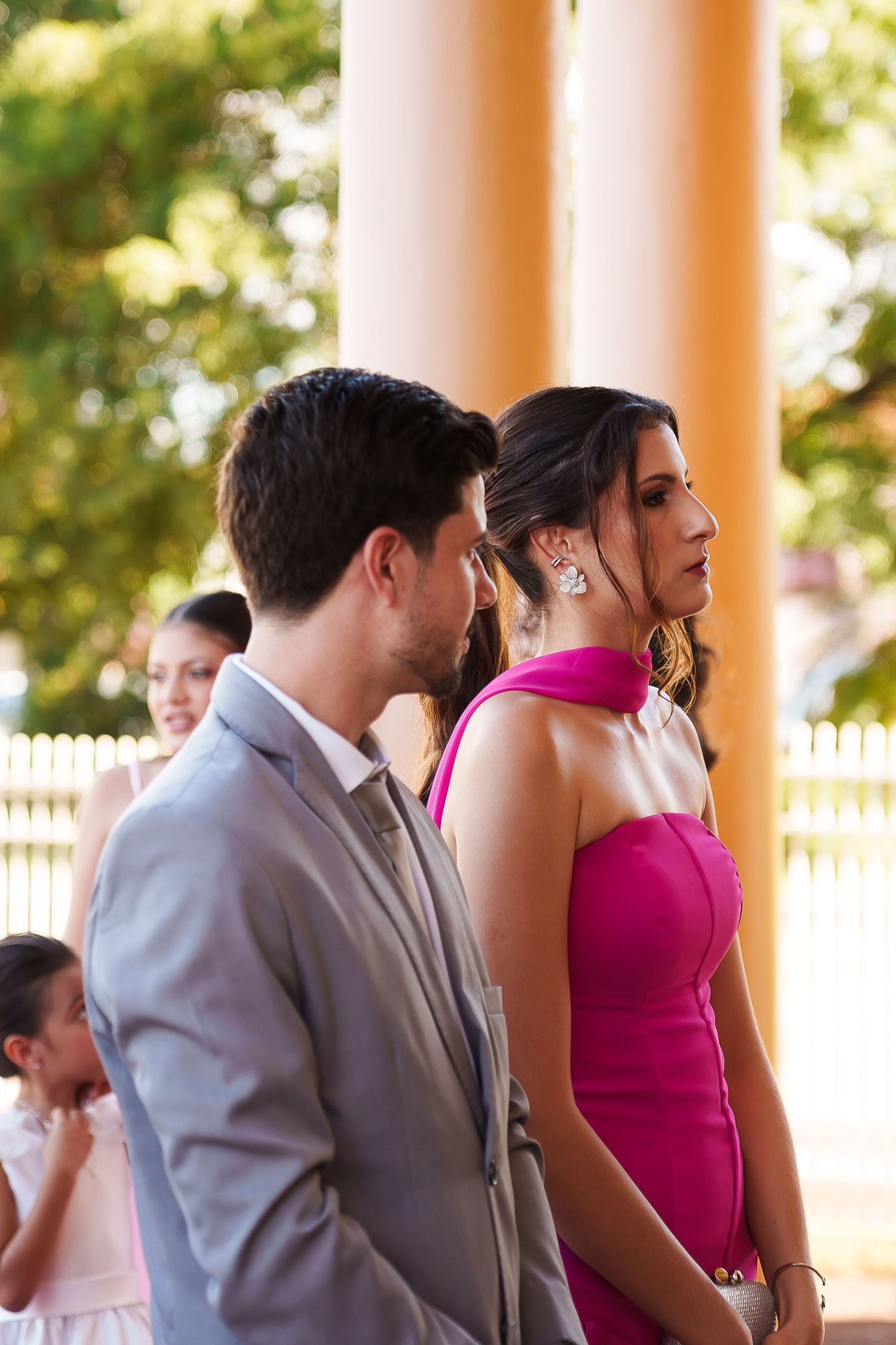 Elegant couple in formal attire, man in gray suit and woman in pink dress, standing at an outdoor wedding ceremony.
