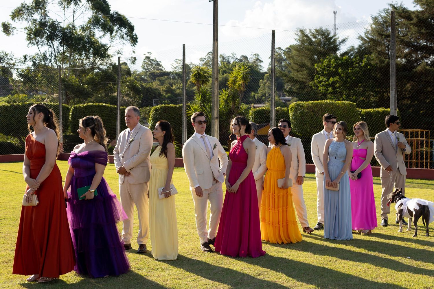 Elegantly dressed group in colorful formal gowns and suits standing on green lawn at outdoor wedding ceremony.