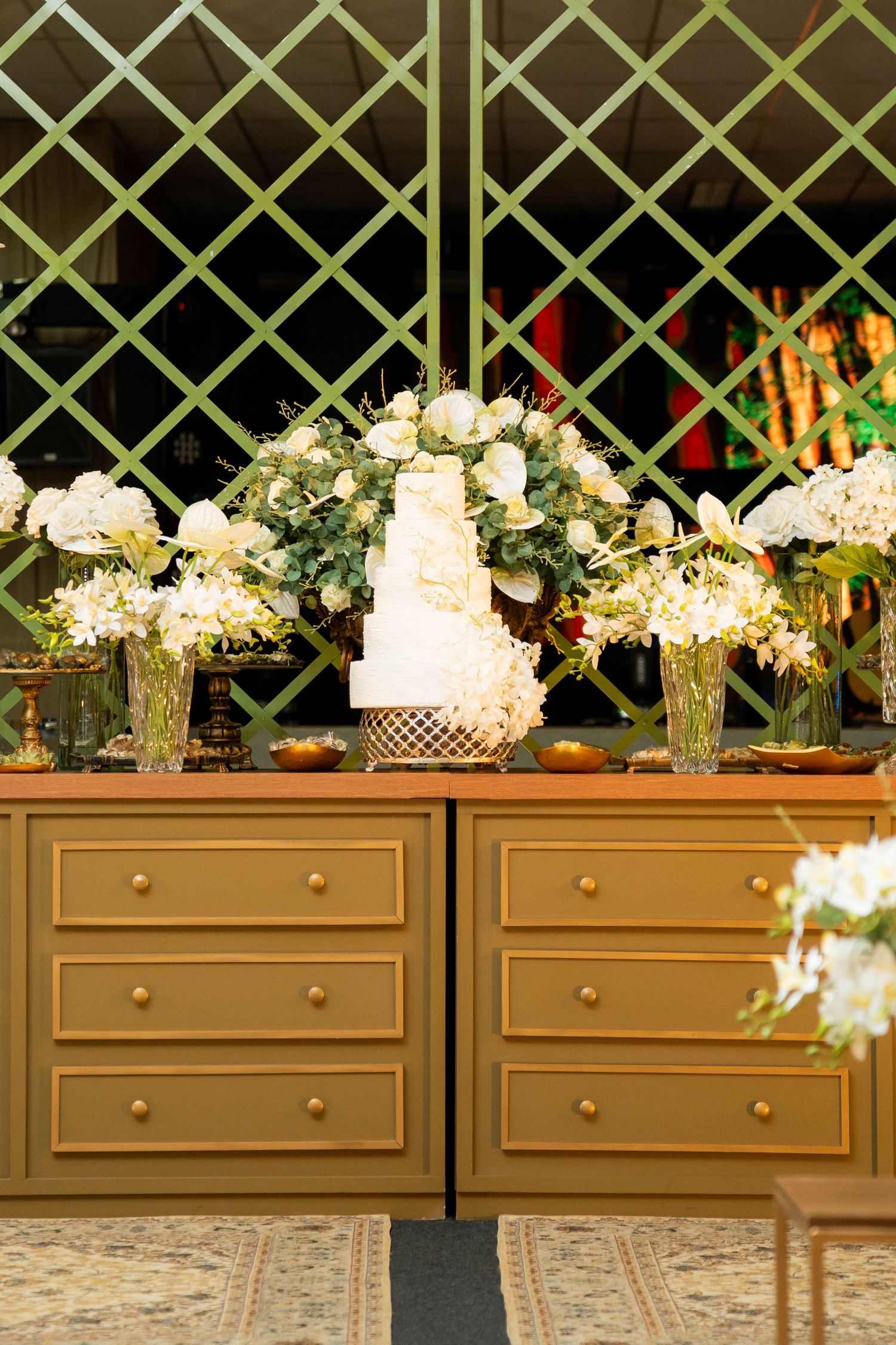 Elegant wedding cake display on gold dresser with white floral arrangements and green lattice backdrop.