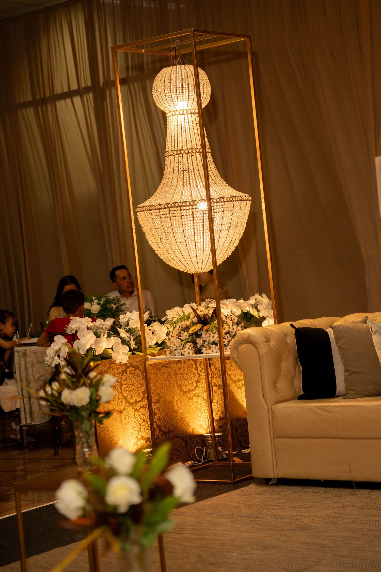 Elegant wedding reception with glowing chandelier, white floral arrangements, and a cream tufted sofa in warm ambient lighting.