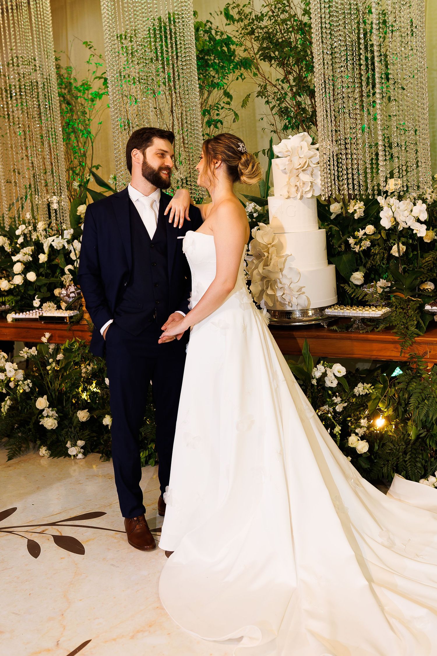 Bride in white strapless gown and groom in navy suit stand together smiling before elegant floral backdrop and tiered wedding cake.