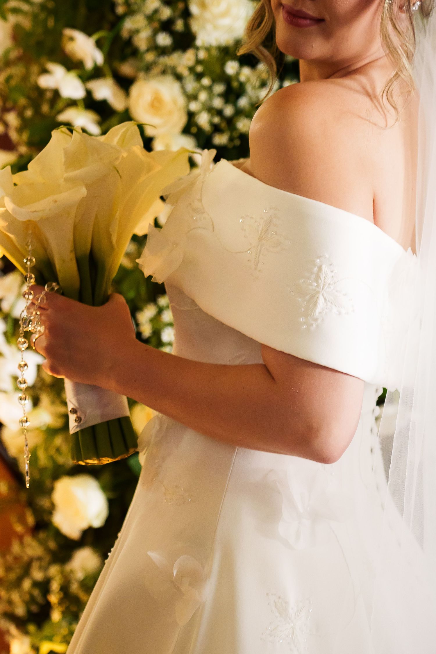 Bride in off-shoulder white gown holding yellow calla lily bouquet with pearl details, standing before floral backdrop.