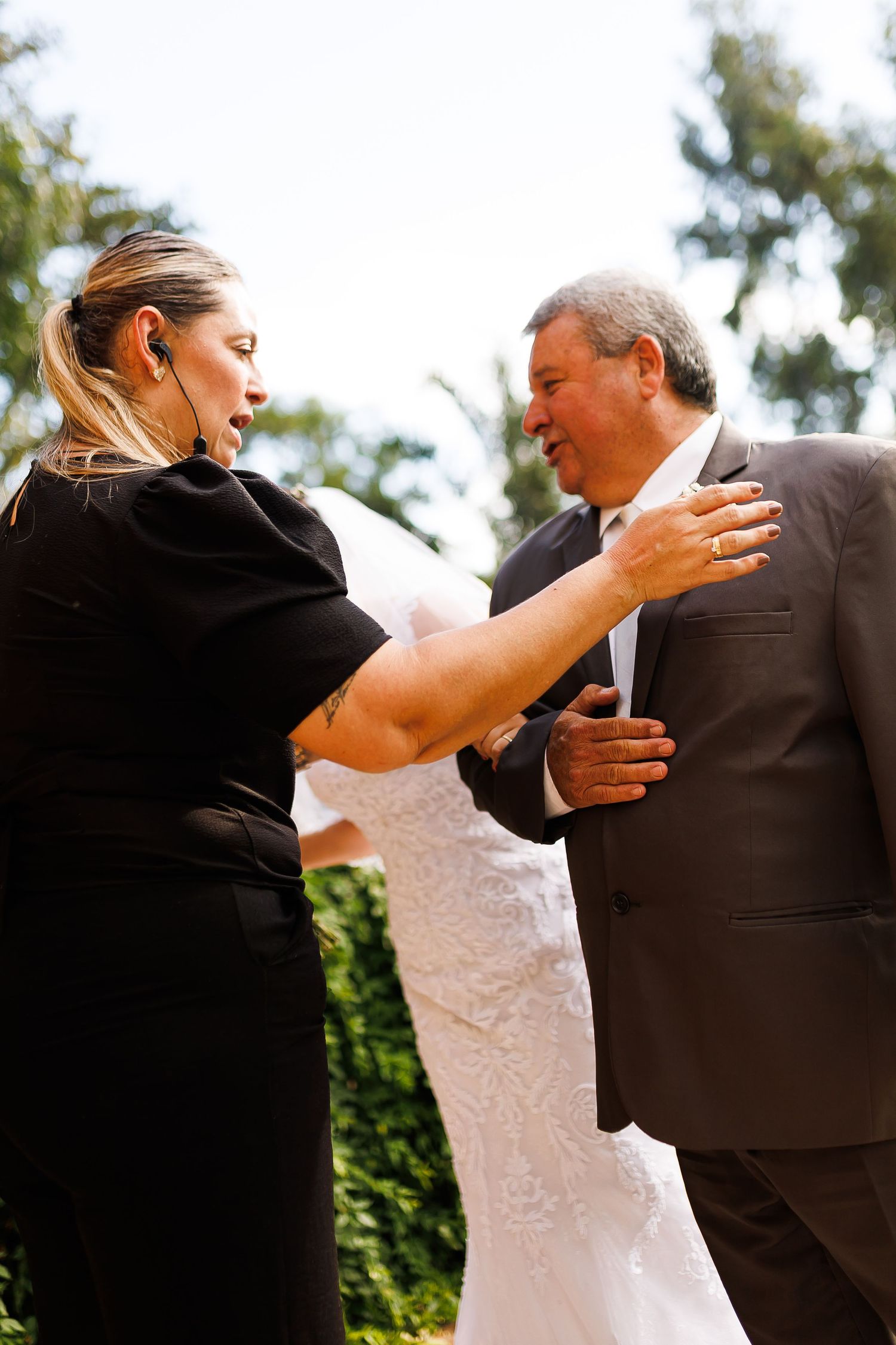 Woman in black dress adjusts mans suit jacket at outdoor wedding ceremony with bride in white visible behind them.