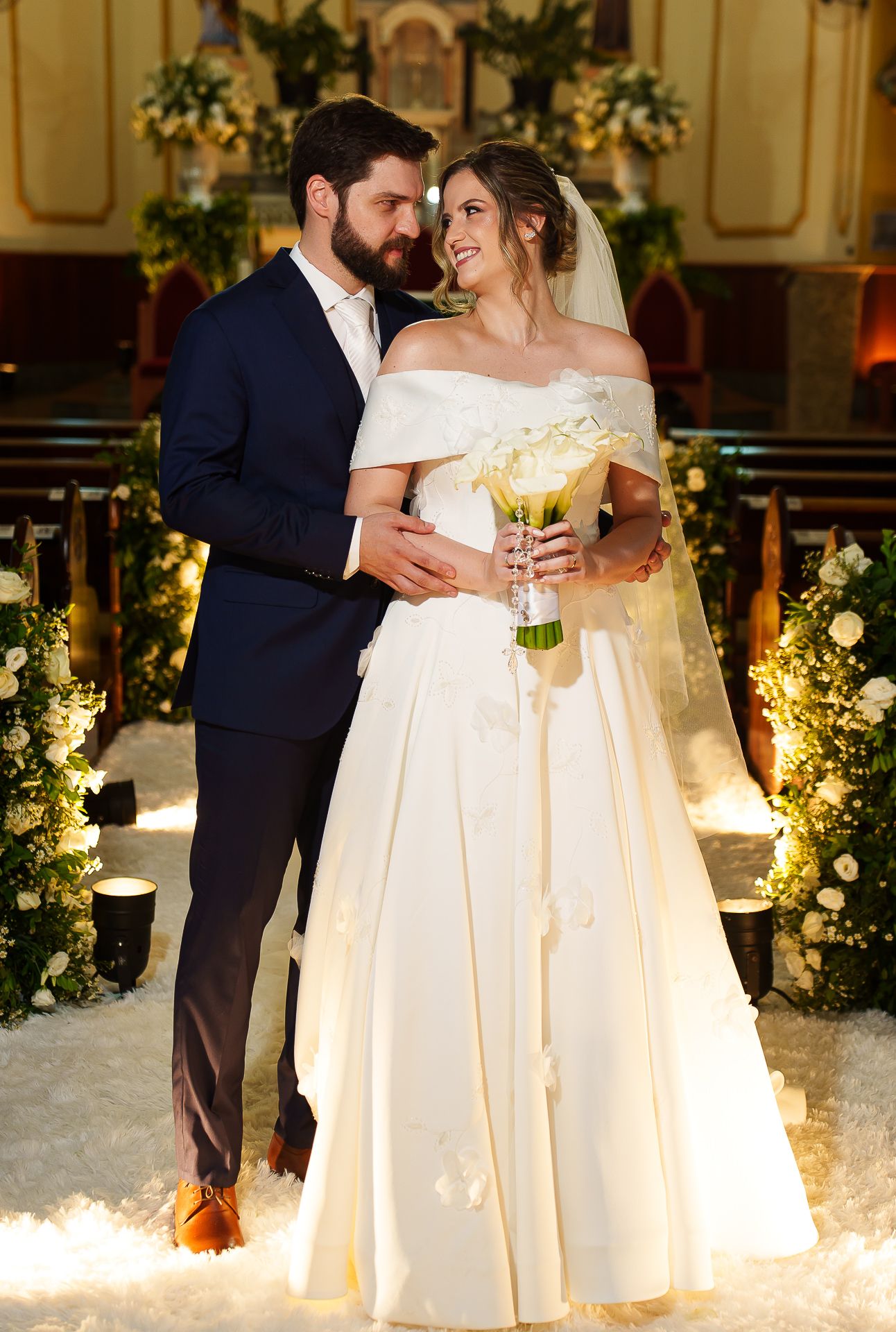 Bride and groom pose together in a church aisle, she holds white roses bouquet wearing off-shoulder gown, he wears navy suit.