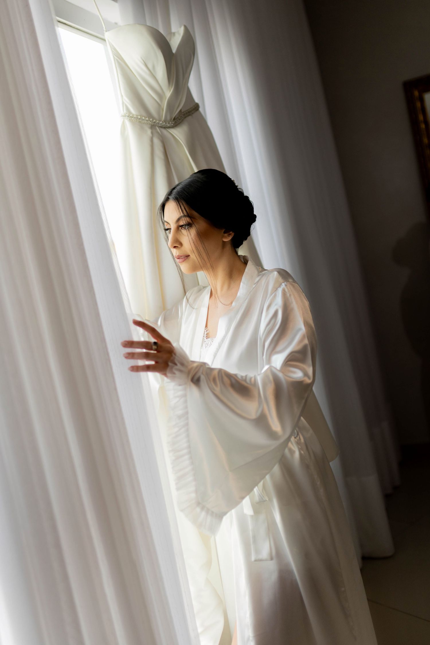Bride in white silk robe gazing at her wedding dress hanging by a sunlit window on her wedding morning.