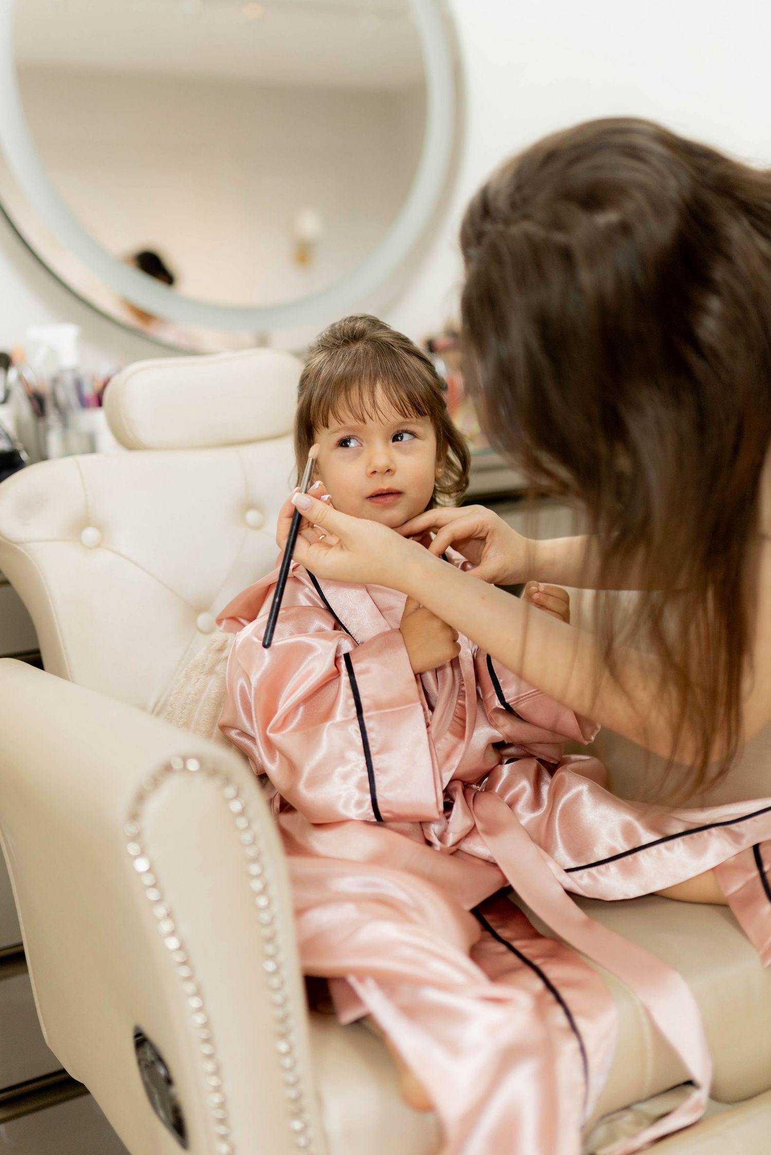A woman in pink satin pajamas helps a little girl dress up in a beauty salon chair.