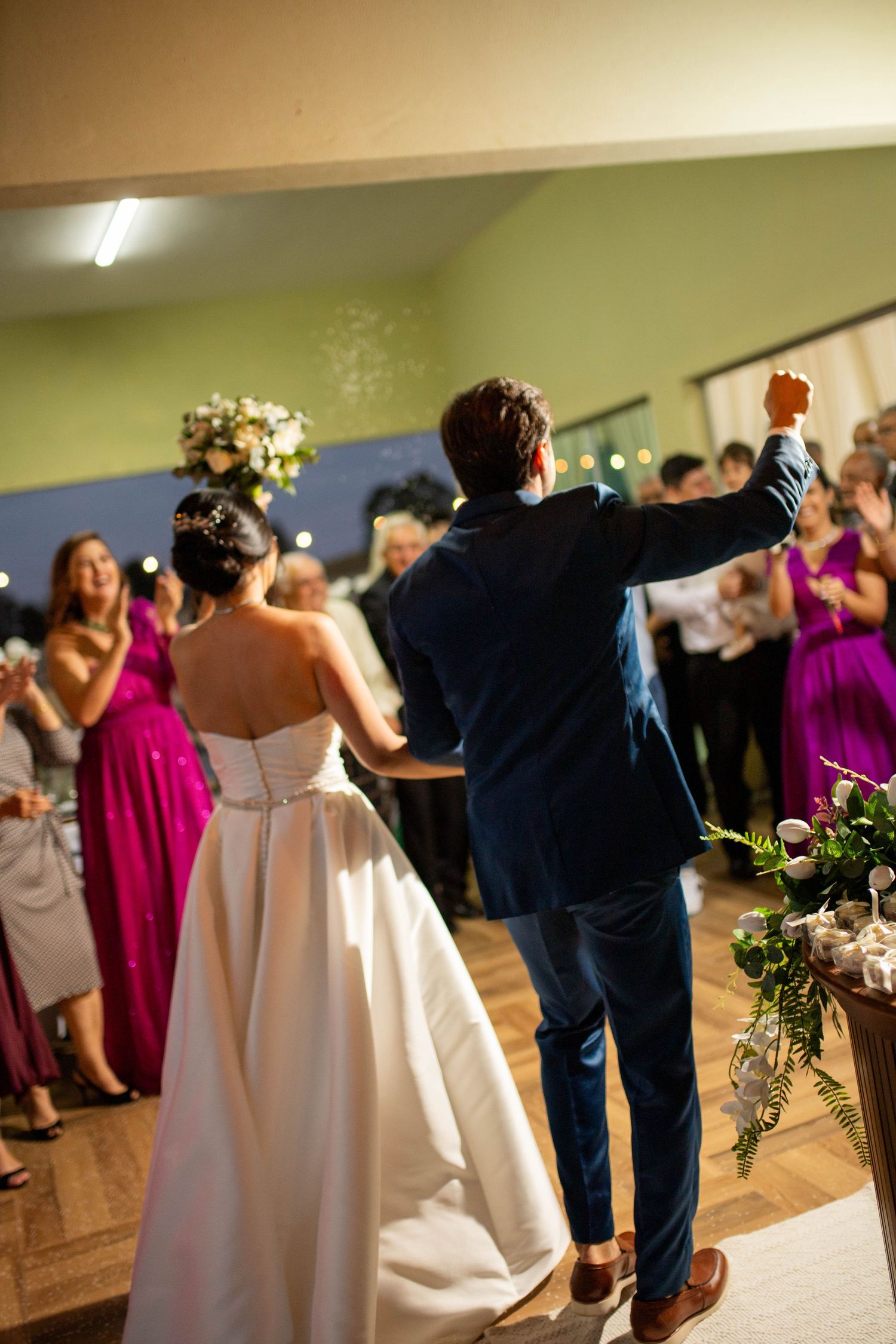 Bride and groom entering wedding reception hall, guests in magenta dresses cheering, wooden dance floor.