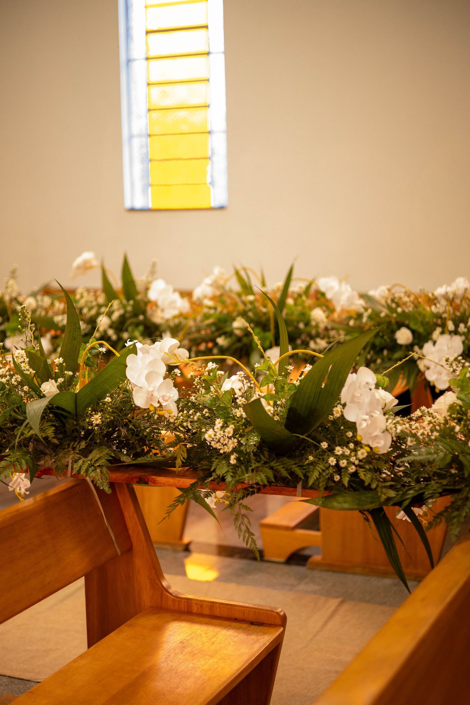 Church pews adorned with lush white floral arrangements and greenery, illuminated by a warm stained glass window.