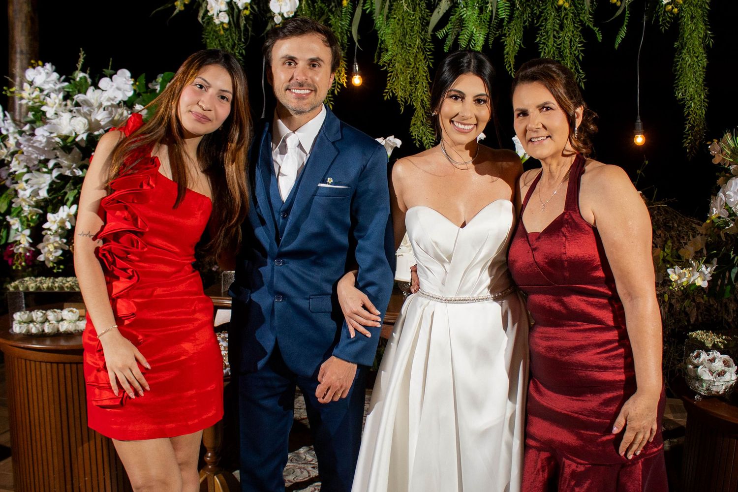 A bride in white gown poses with groom in navy suit and two women in red and burgundy dresses at elegant wedding reception.