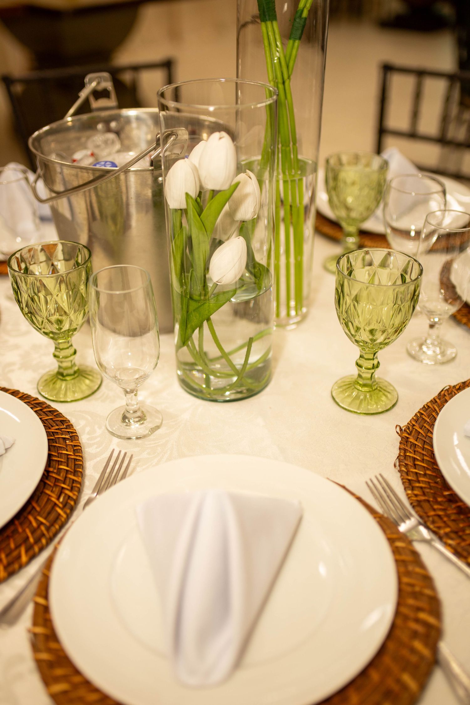 Elegant table setting with white tulips, green goblets, wicker chargers, and white plates with folded napkins.
