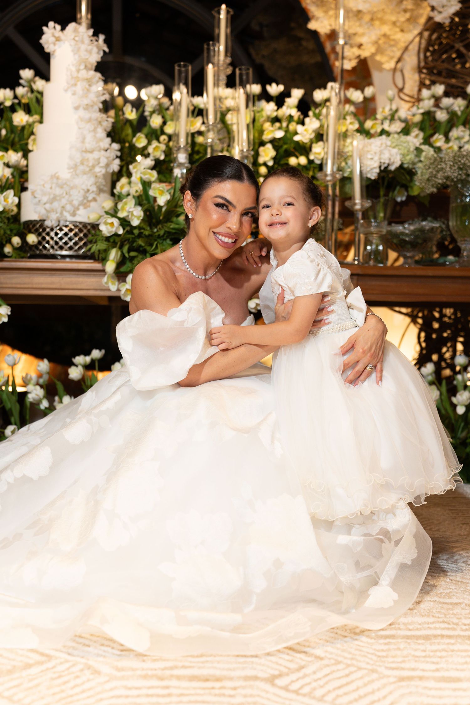 Bride in white ball gown poses with flower girl in matching white dress at elegant wedding reception.